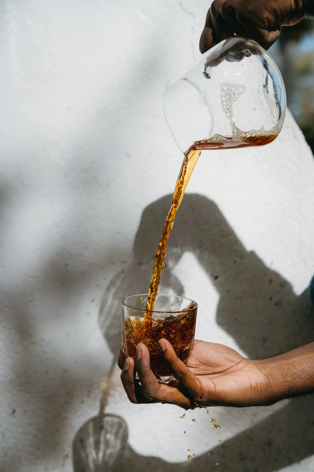 A person is pouring a dark beverage from a glass into a smaller glass held with their hand, with a plain light background.