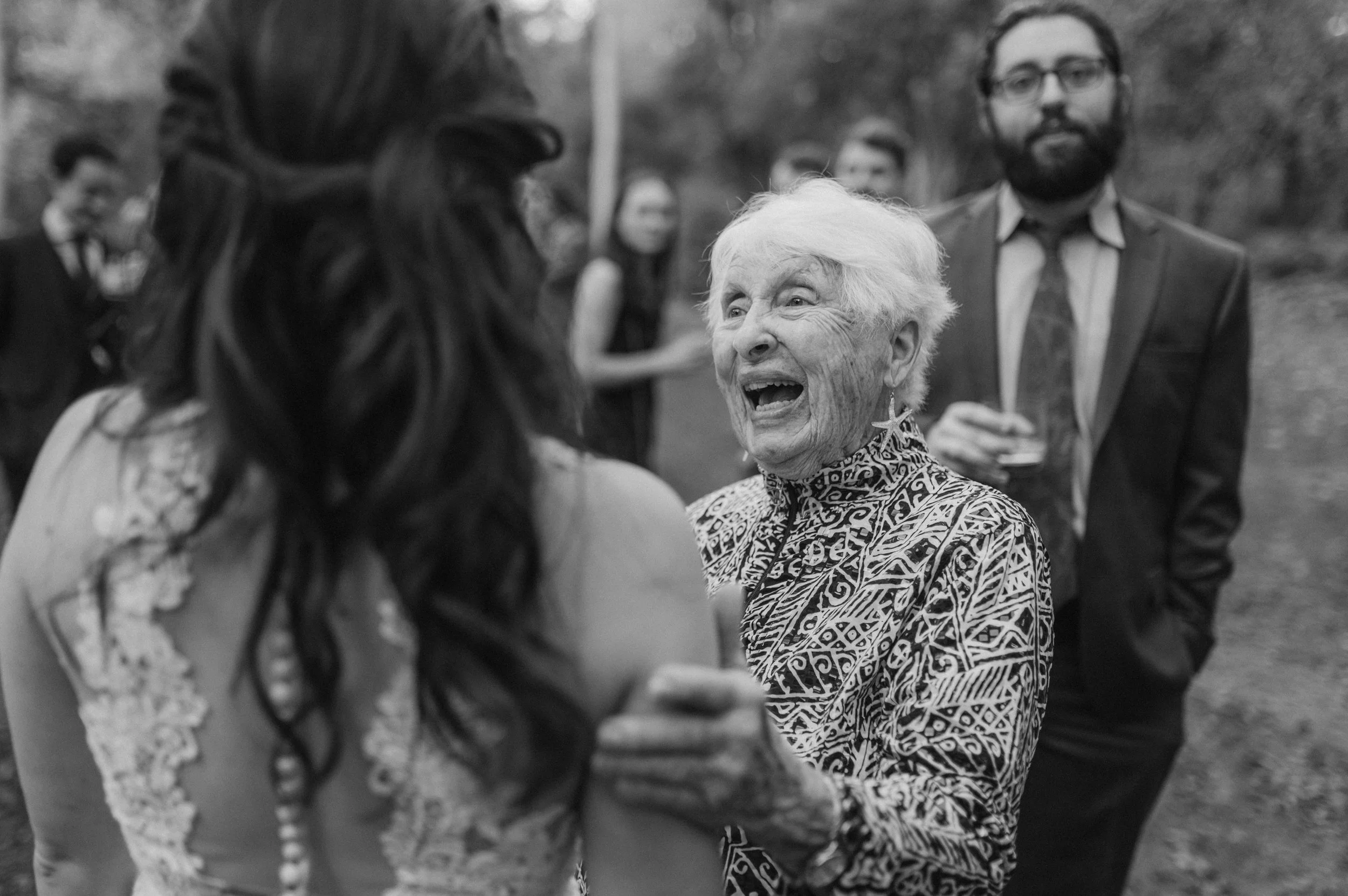 An elderly woman with white hair and tribal patterned clothing is smiling and laughing while holding hands with a younger woman with long, dark hair. In the background, there are several people in formal attire at an outdoor event.