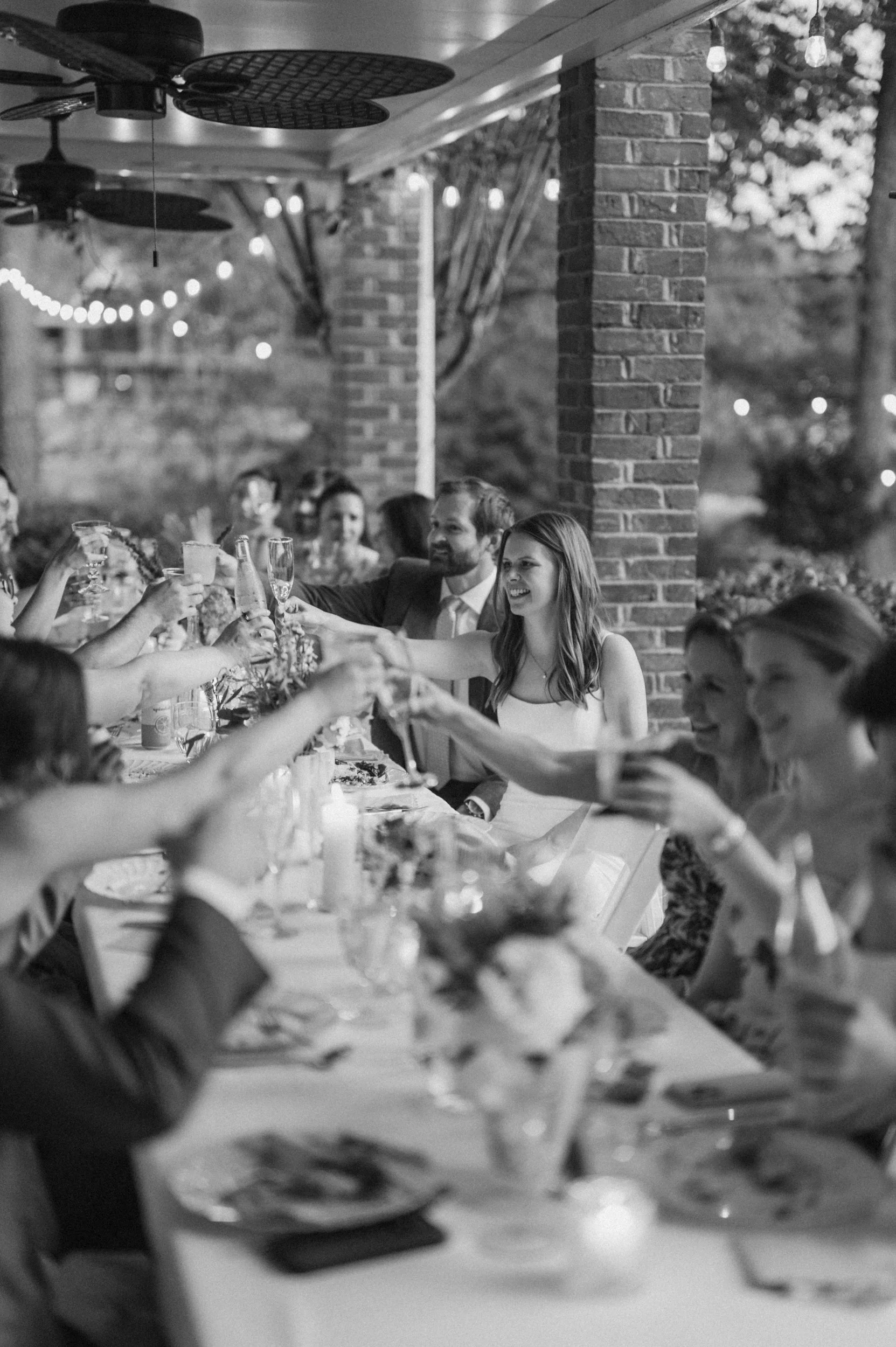 Guests celebrating at a wedding reception, toasting with drinks at a long decorated table outdoors.