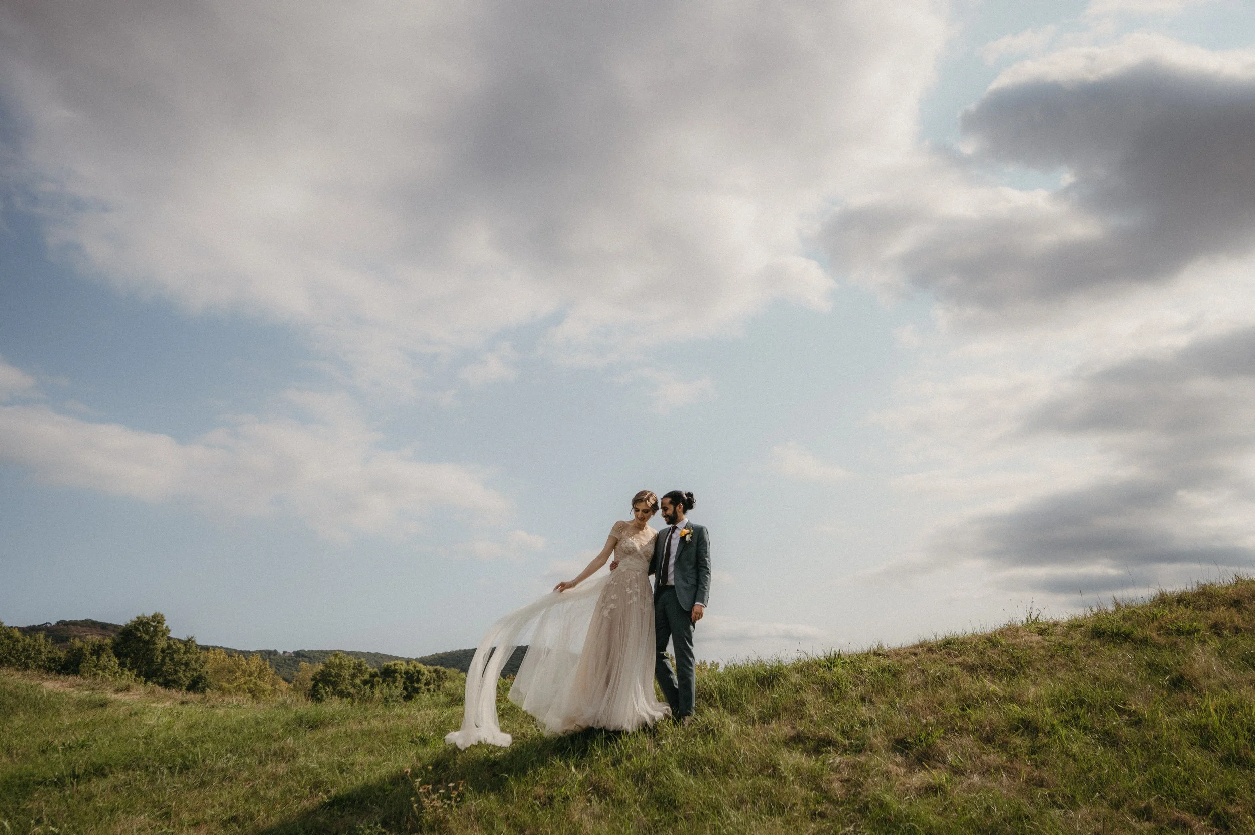 Bride and groom standing together on a grassy hill with a cloudy sky in the background.