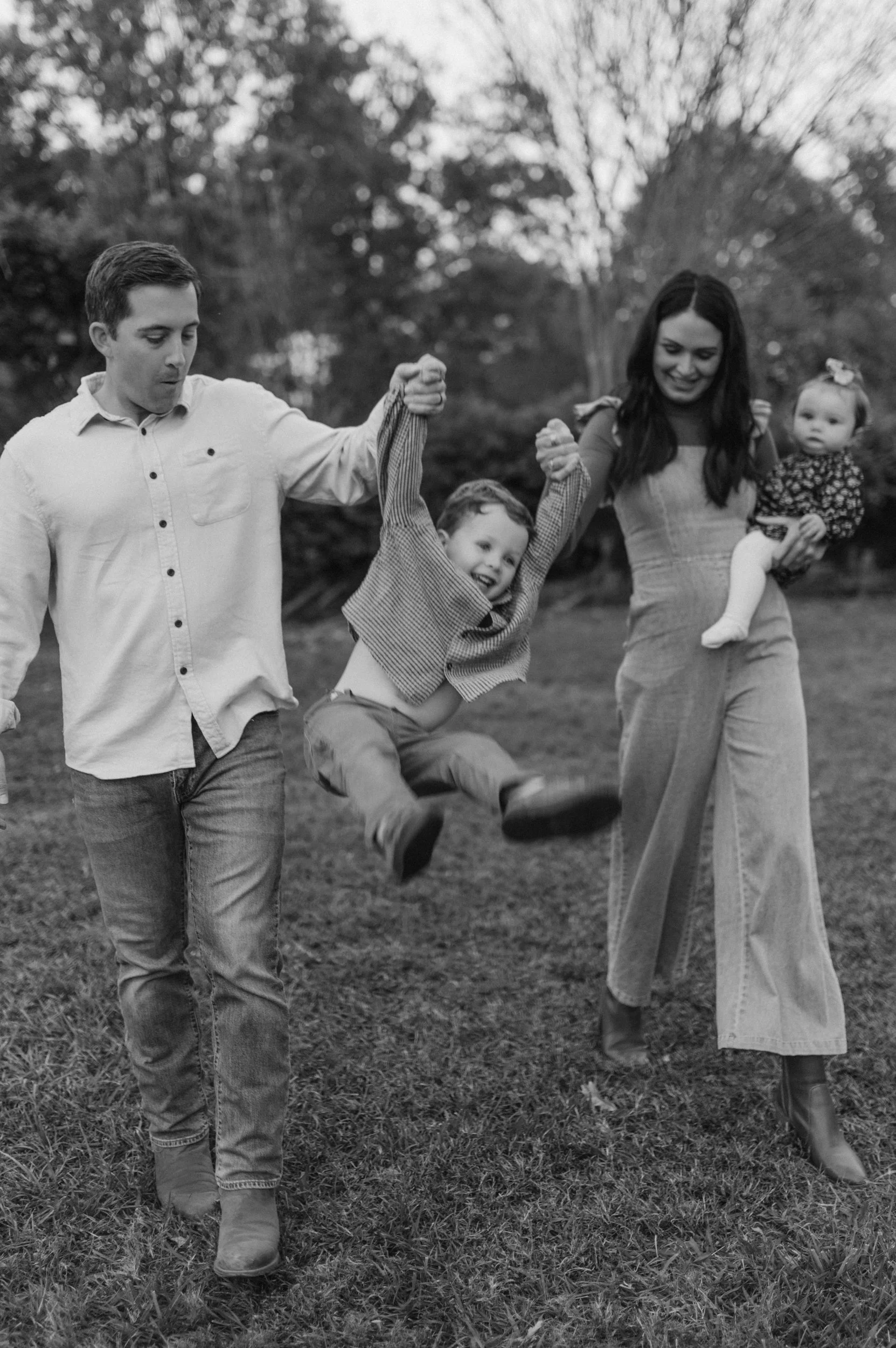 A family of four enjoying an outdoor moment, with a father and mother holding a child’s hands as he swings between them, and the mother holding a baby girl in her arm.