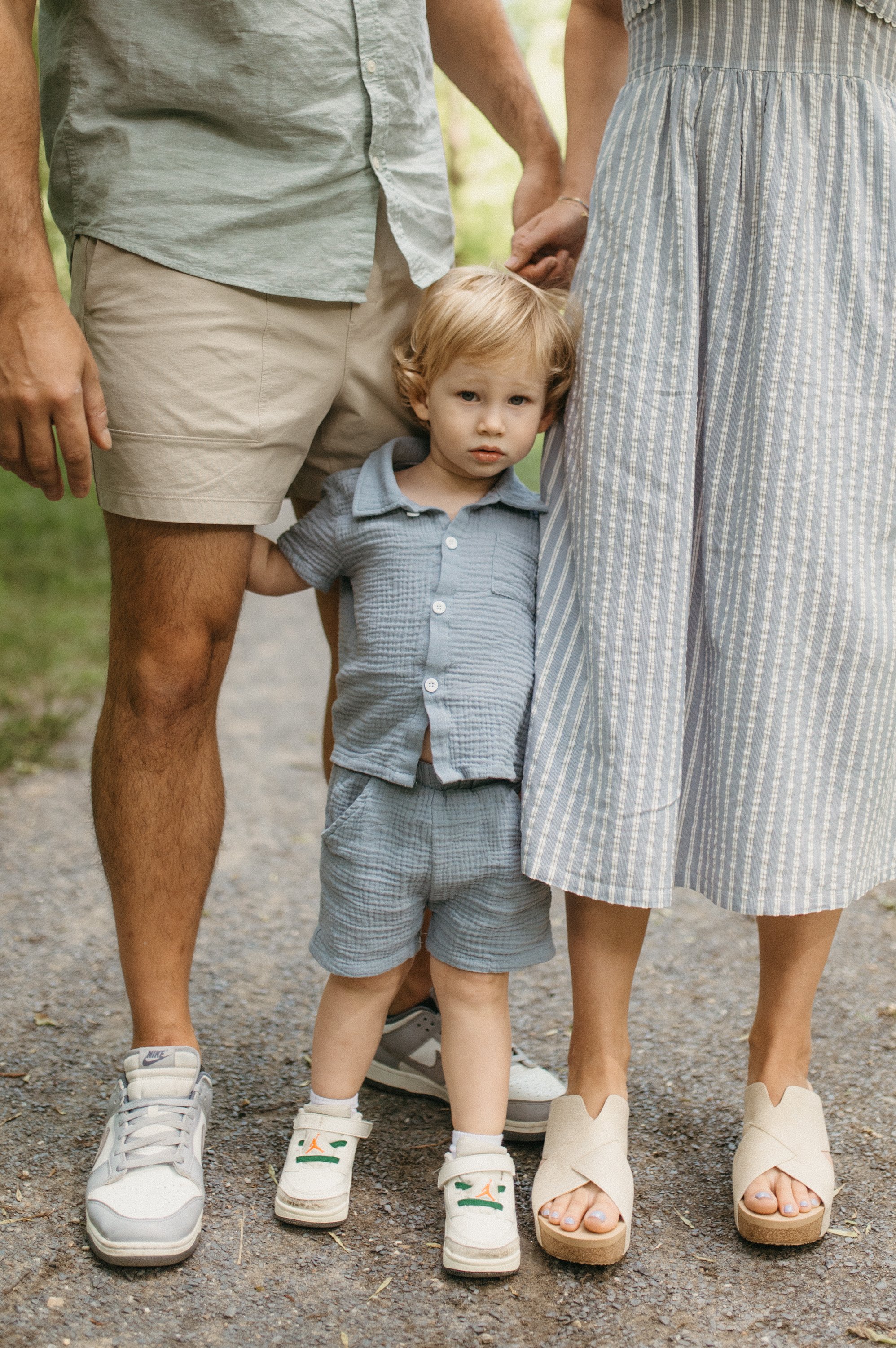 A young boy with blonde hair and a blue outfit standing between two adults, holding their hands. The adults are dressed casually, and only their legs and lower torsos are visible. The scene appears to be outdoors.