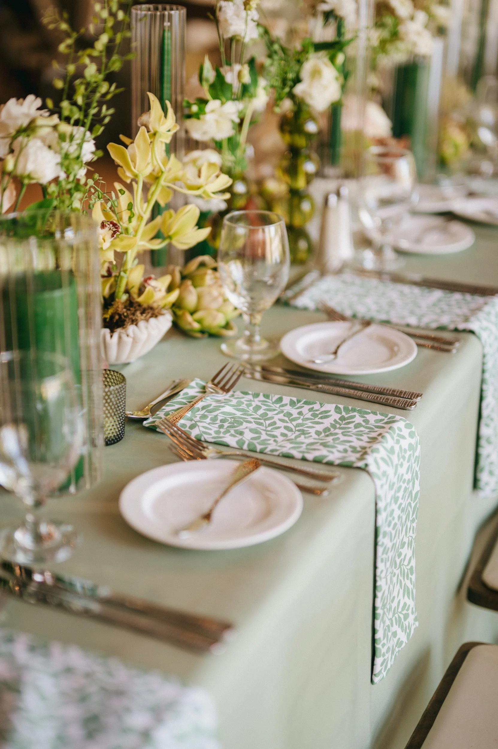 A elegantly set dining table with white plates, silverware, and clear wine glasses. The table is decorated with green and white patterned napkins, floral arrangements in green and white, and tall green candles in glass holders, creating a sophisticat