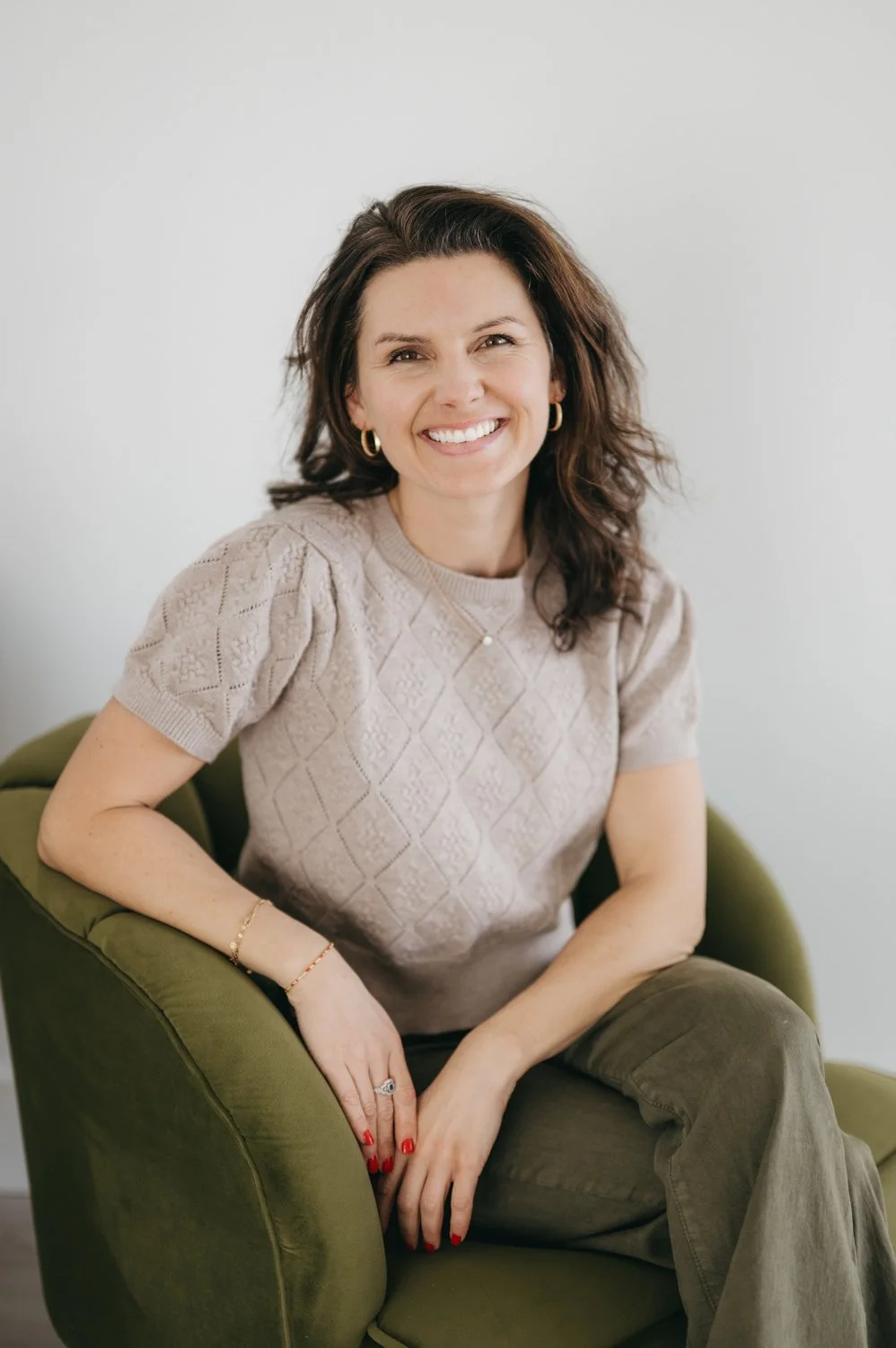 A woman sitting in a green chair, smiling, wearing a beige sweater, and gold jewelry.