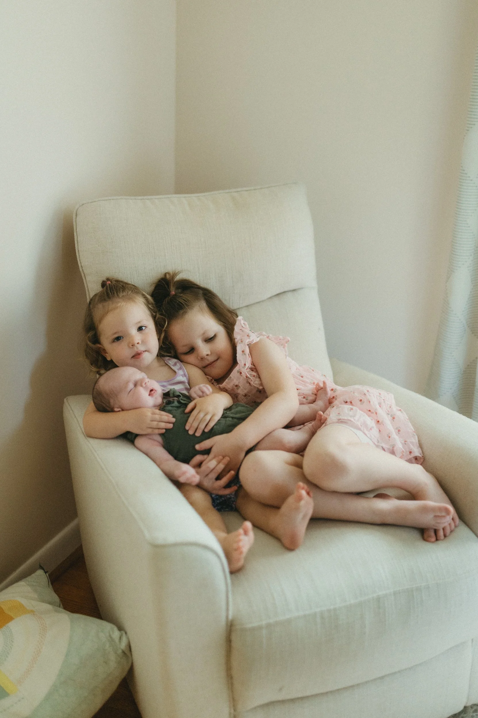 Three young girls sitting on a beige armchair, holding a newborn baby, in a cozy room with neutral walls and a corner window.