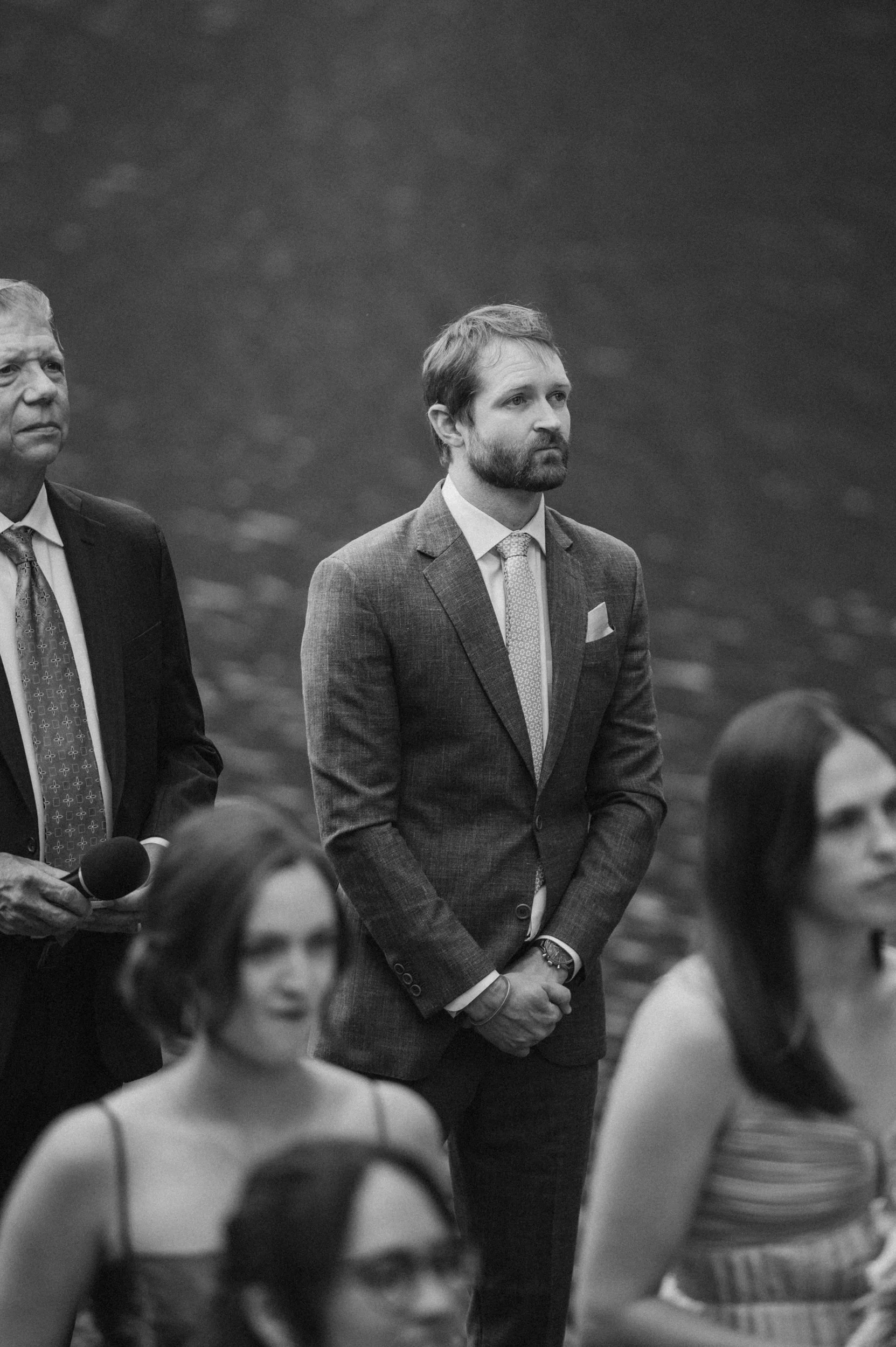 A group of people attending an event, with a man in a suit standing with hands clasped, looking attentive. Others are blurred in the foreground, including a woman and a person holding a microphone.