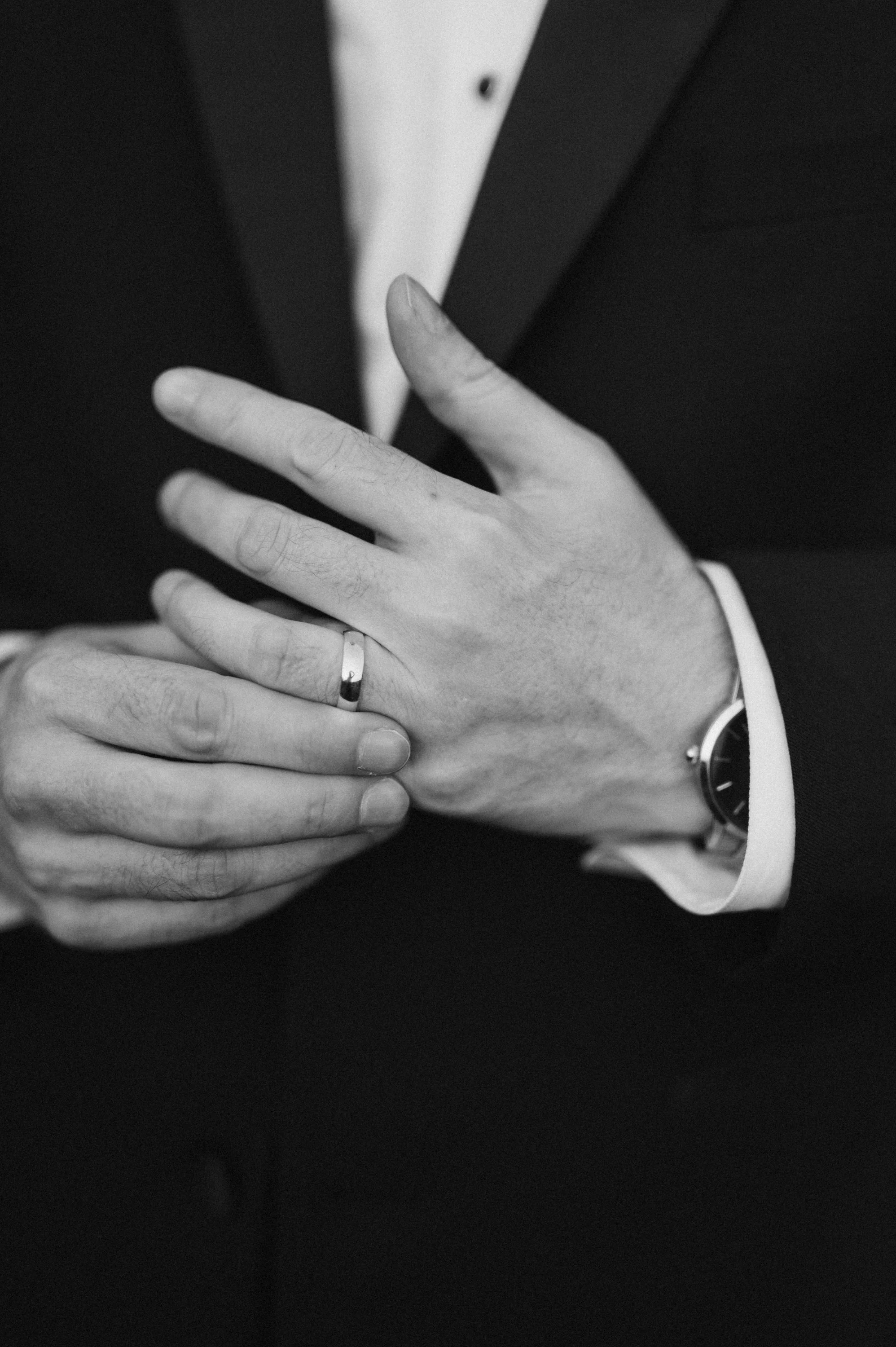 A person in formal attire with a watch on their wrist, adjusting a wedding ring on their finger.