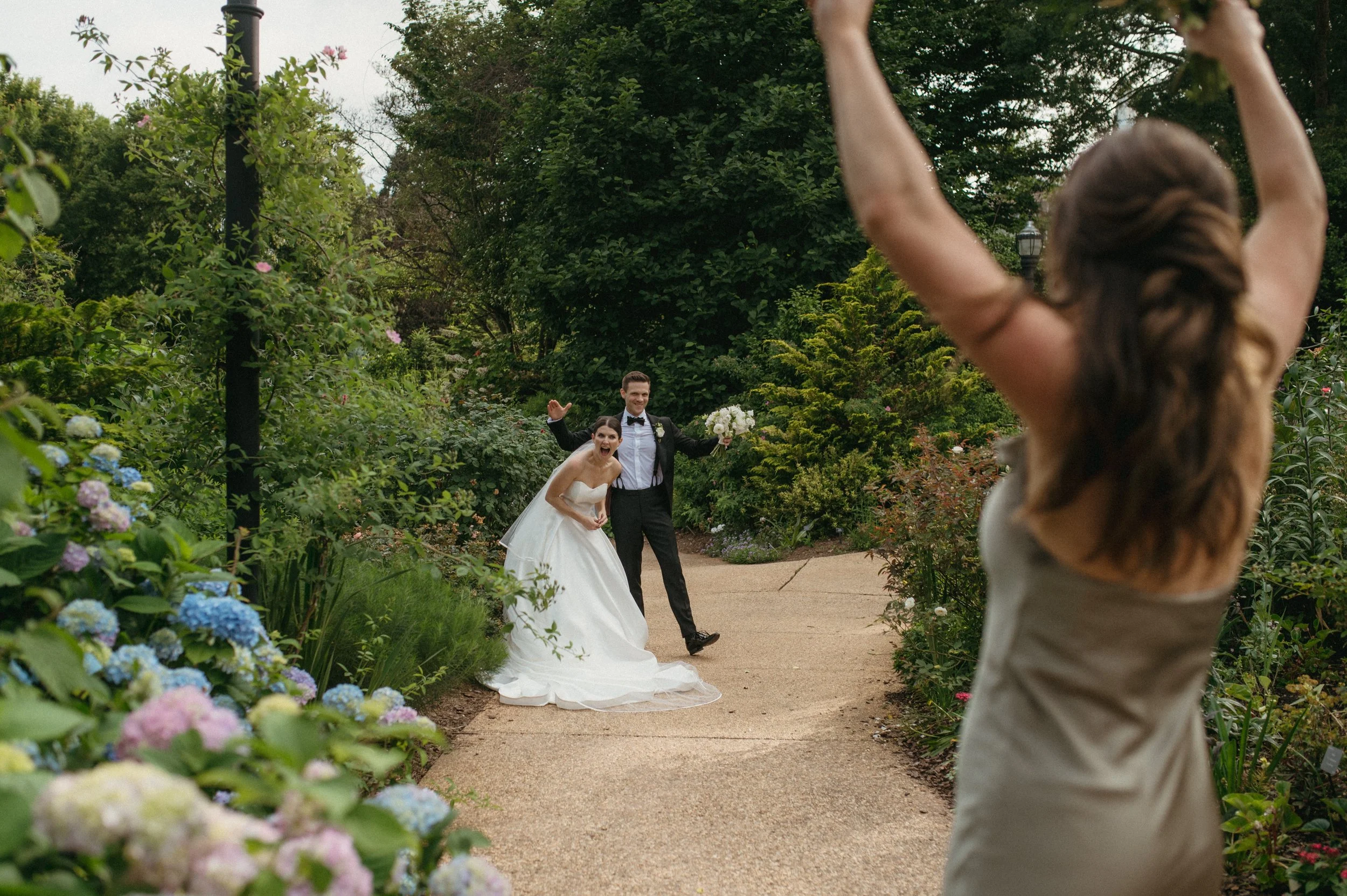 A bride and groom are happily posing for a photo in a lush garden, while a woman with long hair, wearing a sleeveless dress, stands in the foreground with her arms raised. The garden is filled with colorful flowers and greenery.