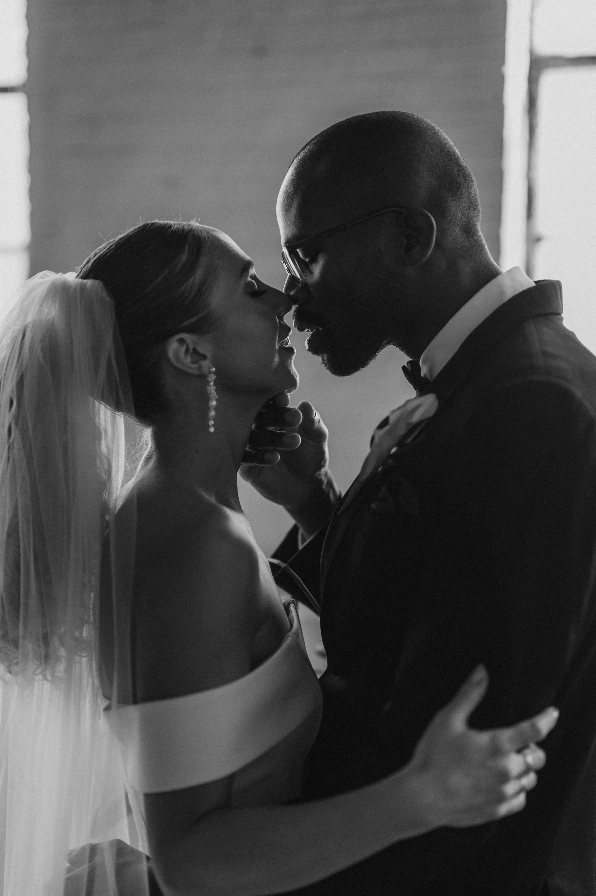 A black and white photograph of a bride and groom close together, about to kiss, with the groom gently holding the bride's chin and her hand on his chest.
