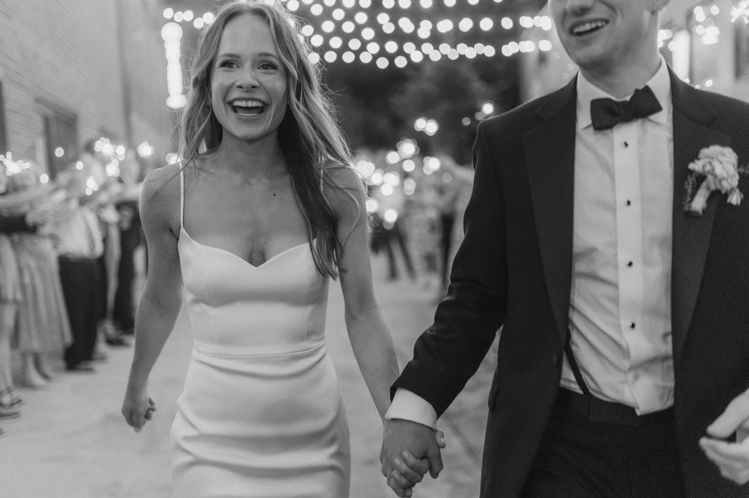 A woman in a white dress and a man in a tuxedo holding hands, smiling at a nighttime celebration or wedding, with string lights overhead and blurred festive decorations in the background.