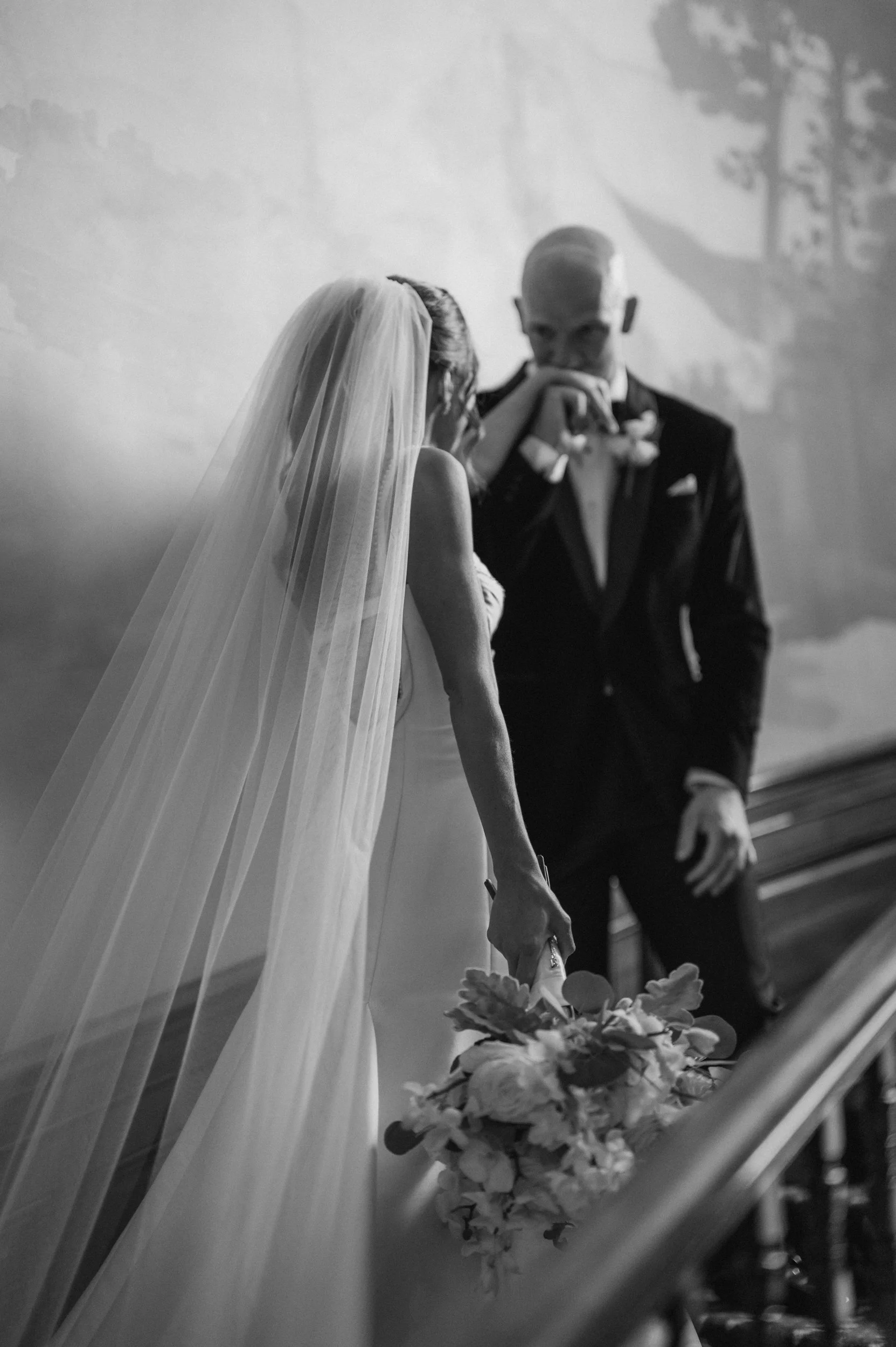 A bride and groom stand apart on a staircase as the groom kisses the top of the brides hand