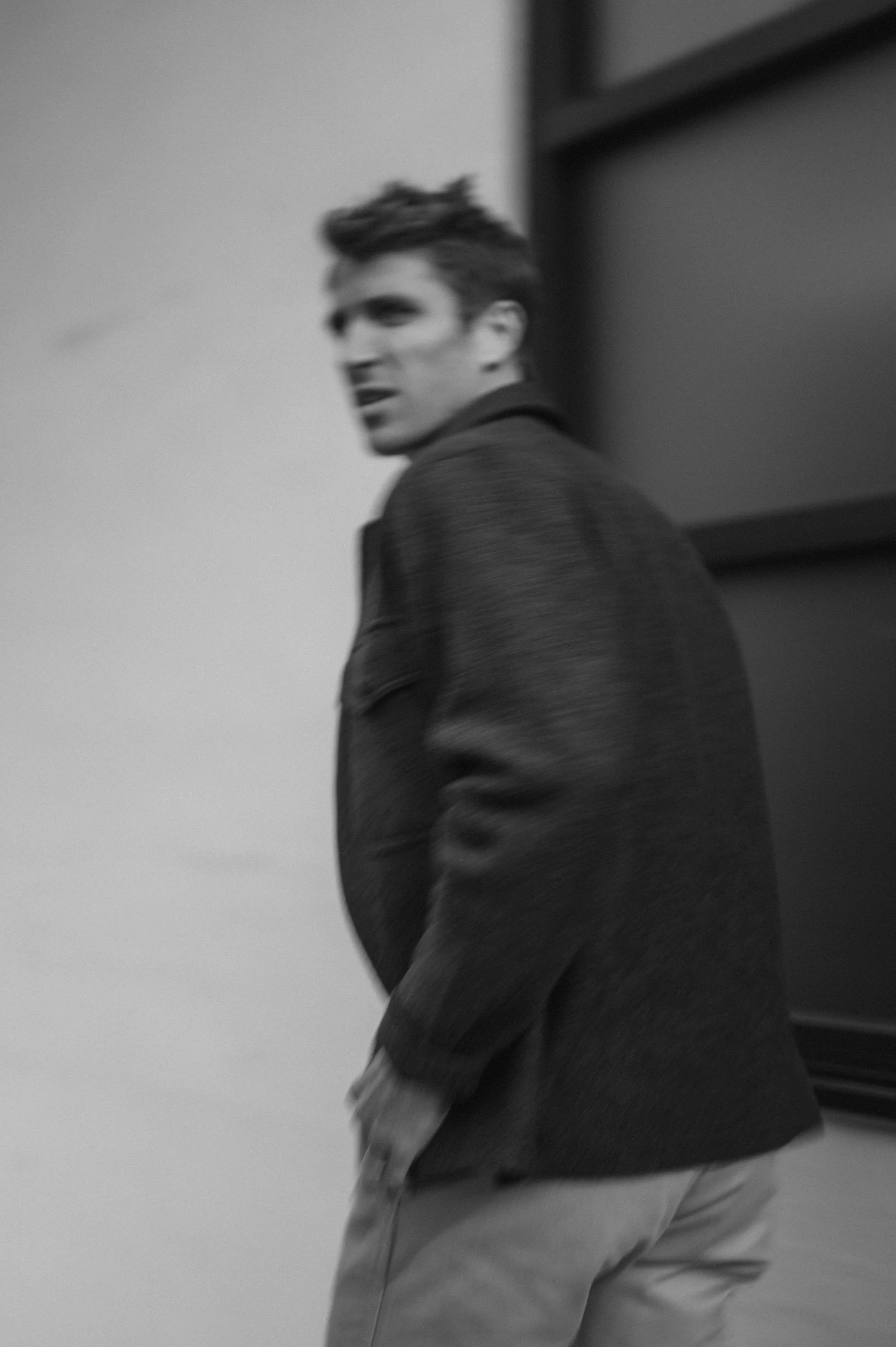 Black and white photo of a young man looking back, wearing a dark jacket and light-colored pants, standing near a wall or safe, with a slightly blurred background.