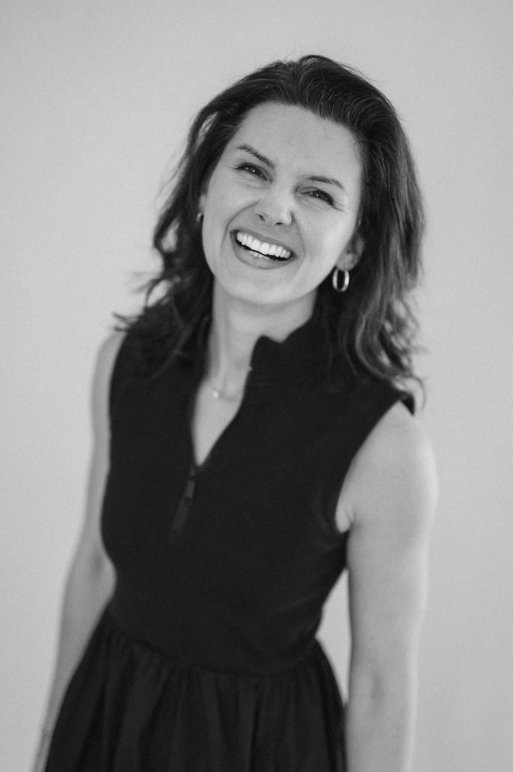 Black and white portrait of a woman with shoulder-length wavy hair, smiling, wearing hoop earrings and a sleeveless zip-up top.