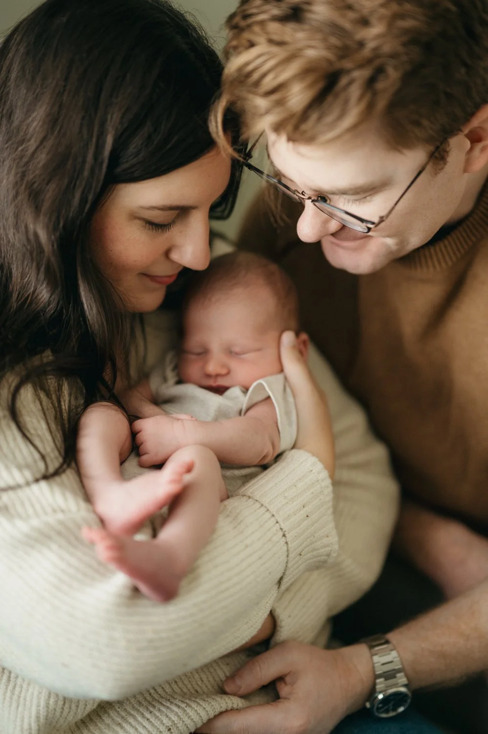 A woman and a man with glasses gently holding and looking at a sleeping newborn baby with closed eyes, in a close, loving, tender moment.