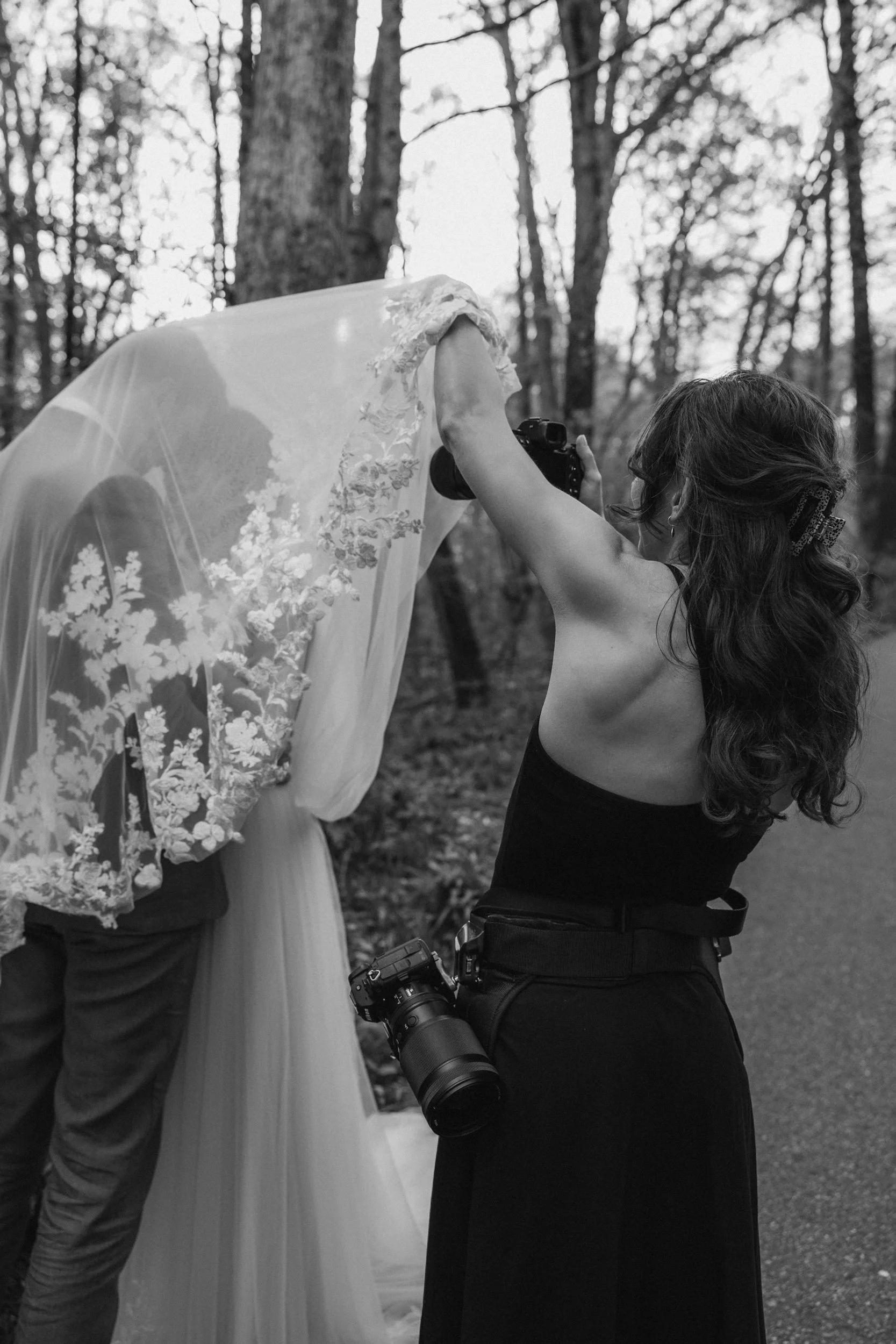 Photographer adjusting a wedding dress on a bride, outdoors in a wooded area, black and white photo.