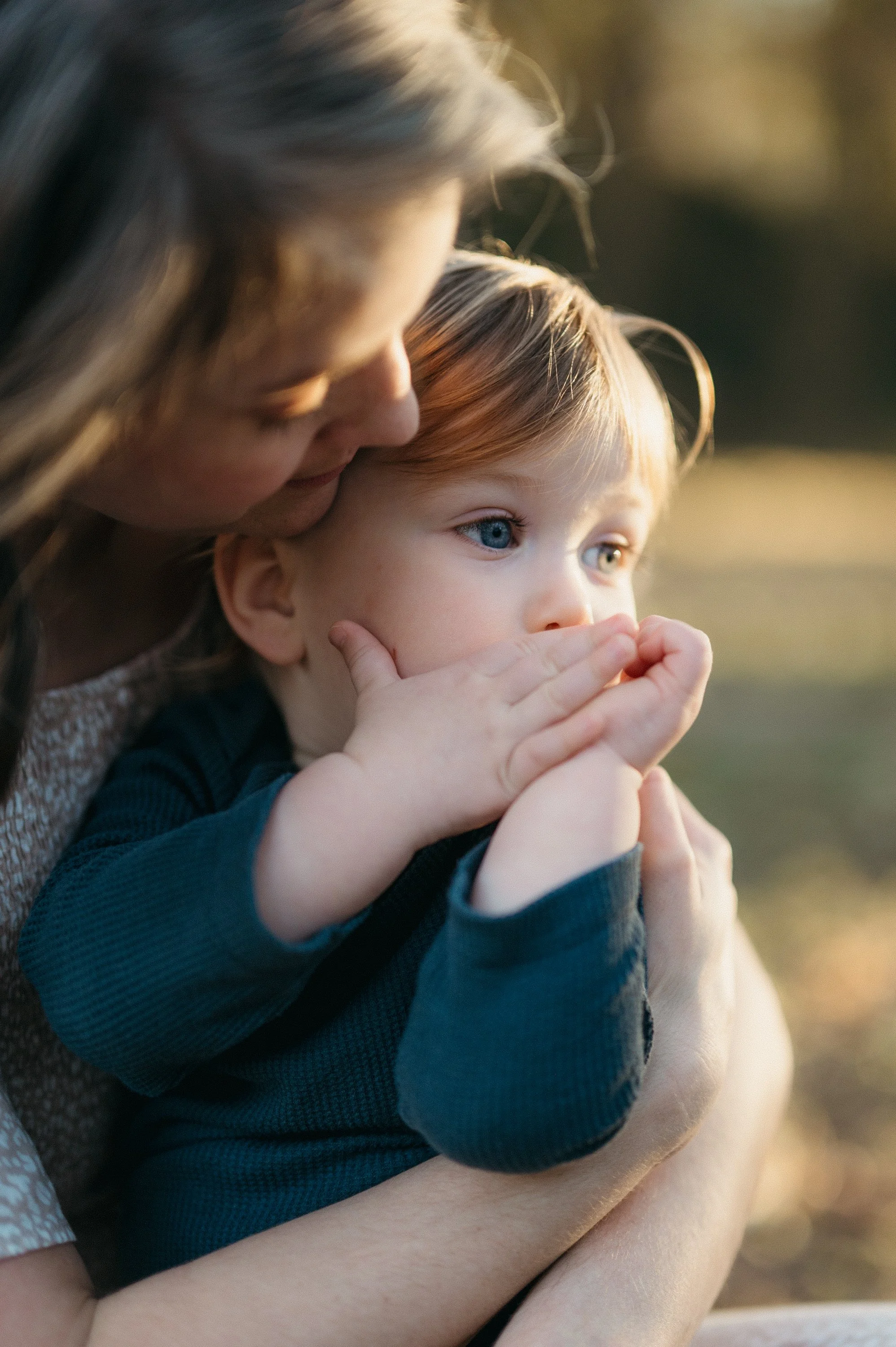 A woman holding a young child with red hair and blue eyes, both outdoors during golden hour, with the woman gently kissing the child's head.