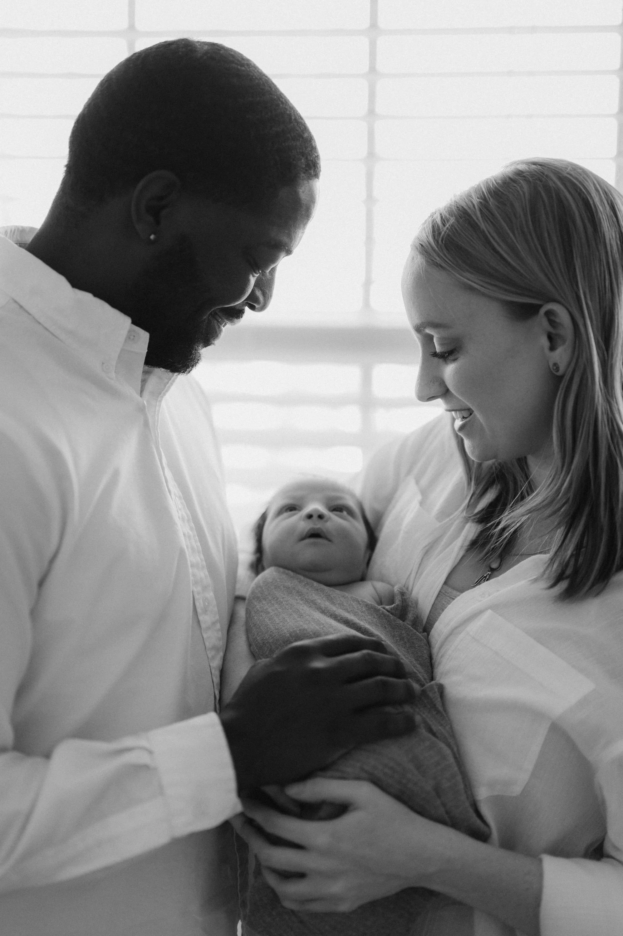 A happy interracial couple holding their newborn baby and gazing at each other in a brightly lit room.