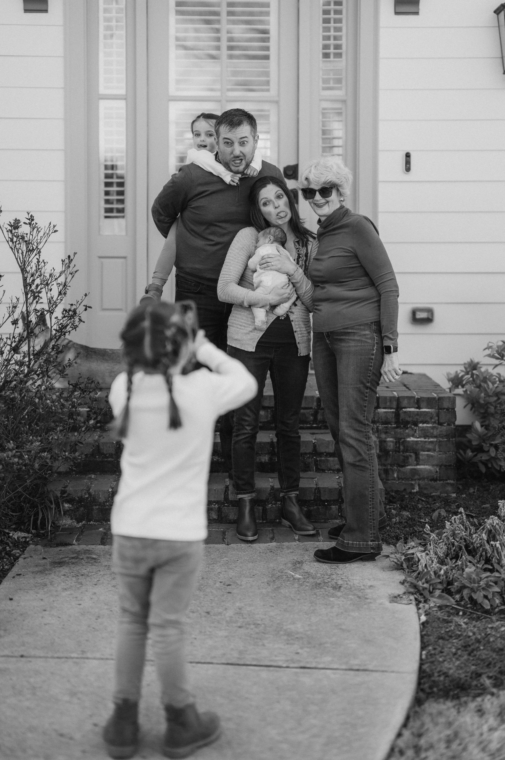 Family photo with five members posing on the front steps of a house, one child taking the picture, and one of the women holding a baby.