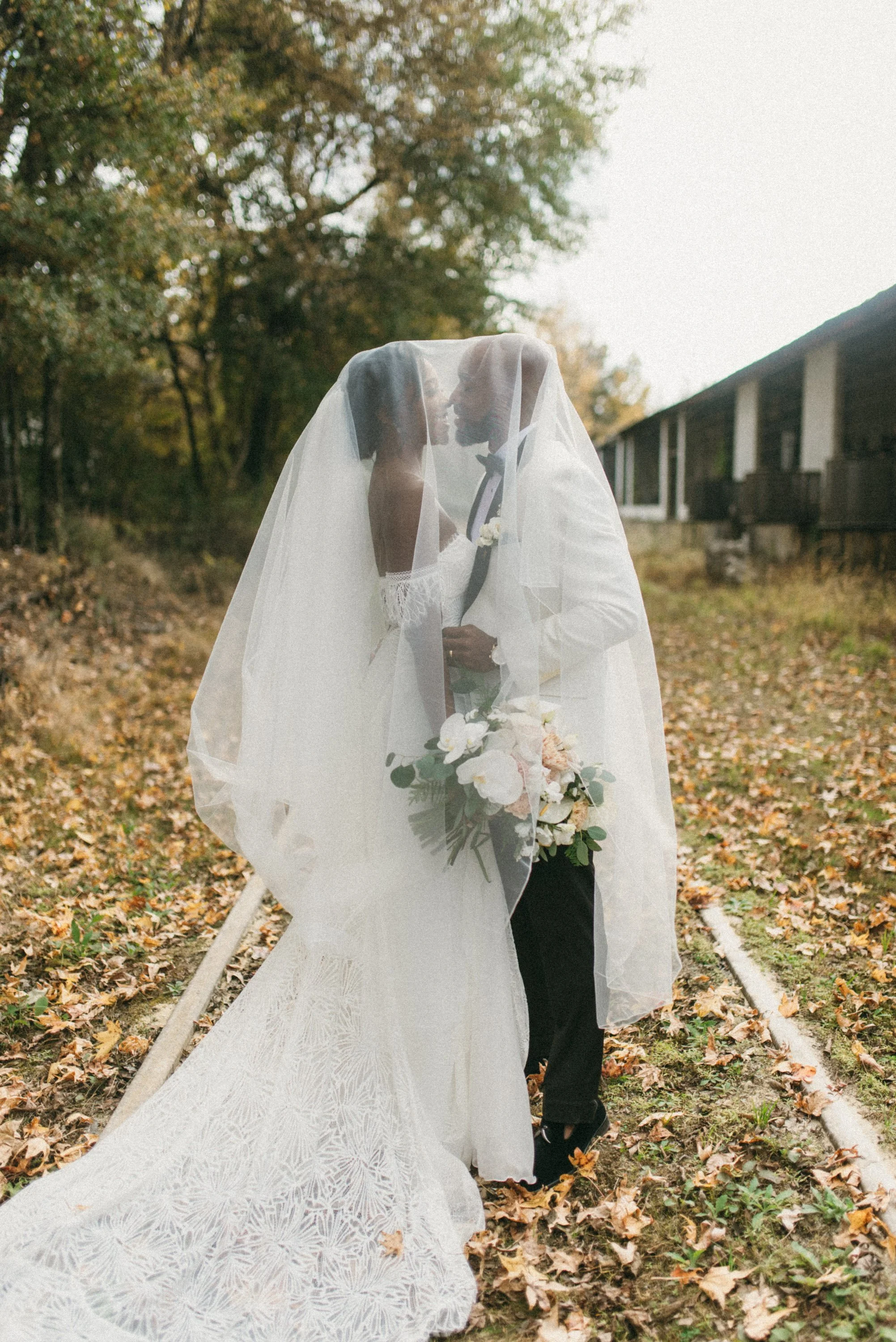 A bride and groom standing close together outdoors, partially covered by a sheer veil, holding a bouquet of white and blush flowers, on a wooded area with autumn leaves.