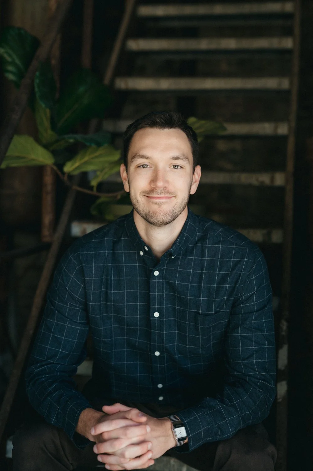A young man with dark hair, light skin, and a beard, smiling, sitting with hands clasped, wearing a checked dark shirt, indoors with stairs and leafy plants in the background.