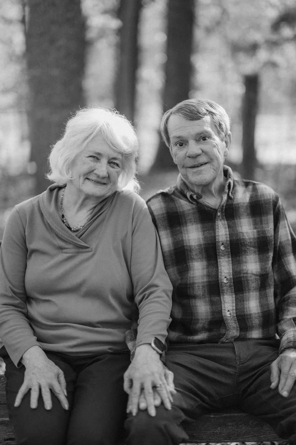 An elderly woman and man sitting outdoors on a bench, smiling at the camera with a blurred forest background.