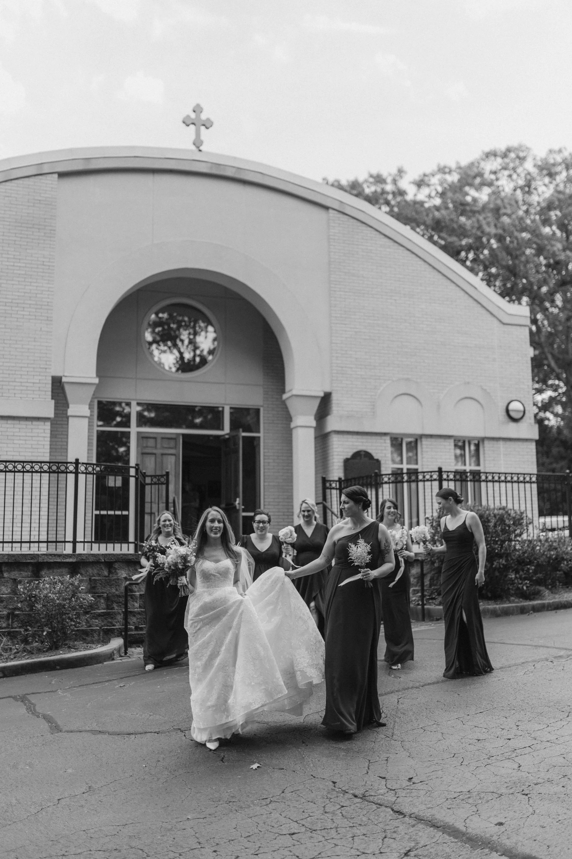 Bride in wedding dress walking outside a church with six women in dark dresses holding bouquets.