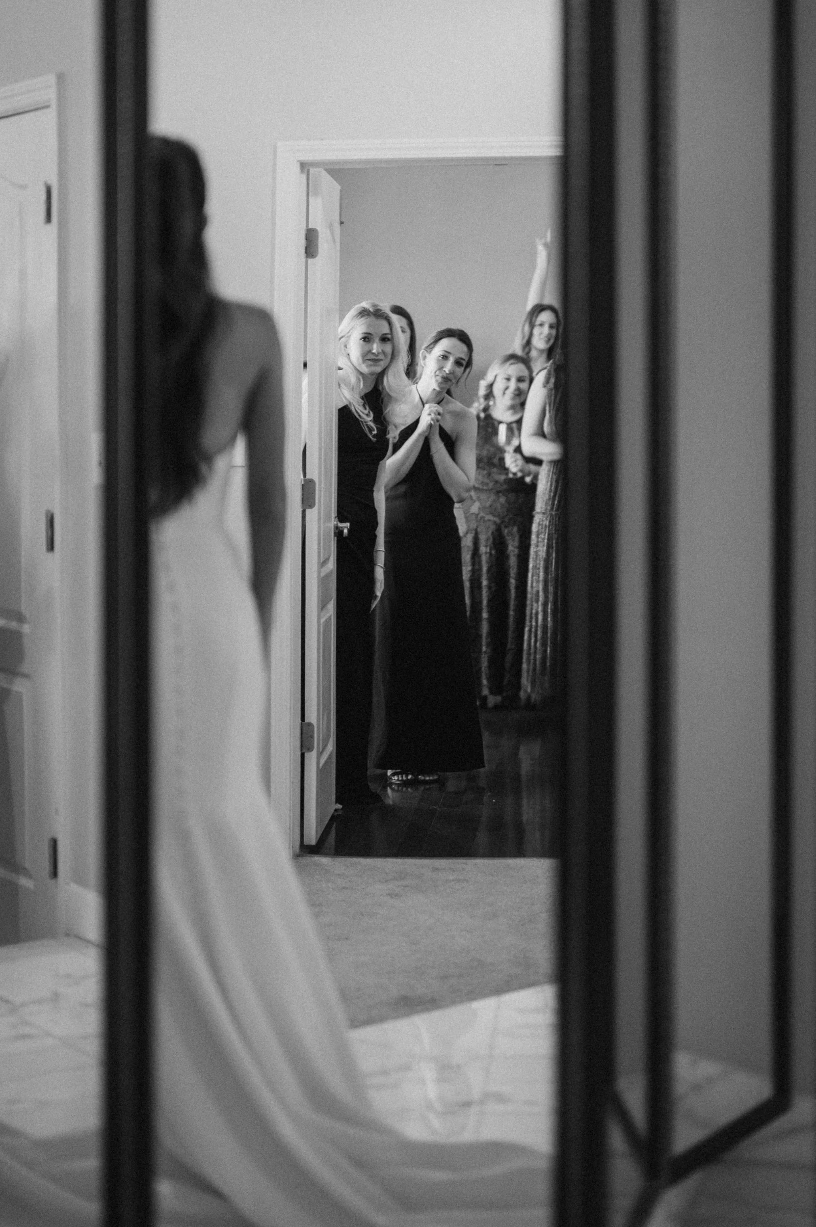 A bride in a wedding dress looking at her reflection in a mirror, with her friends and bridesmaids appearing in the reflection, reacting with joy and admiration.