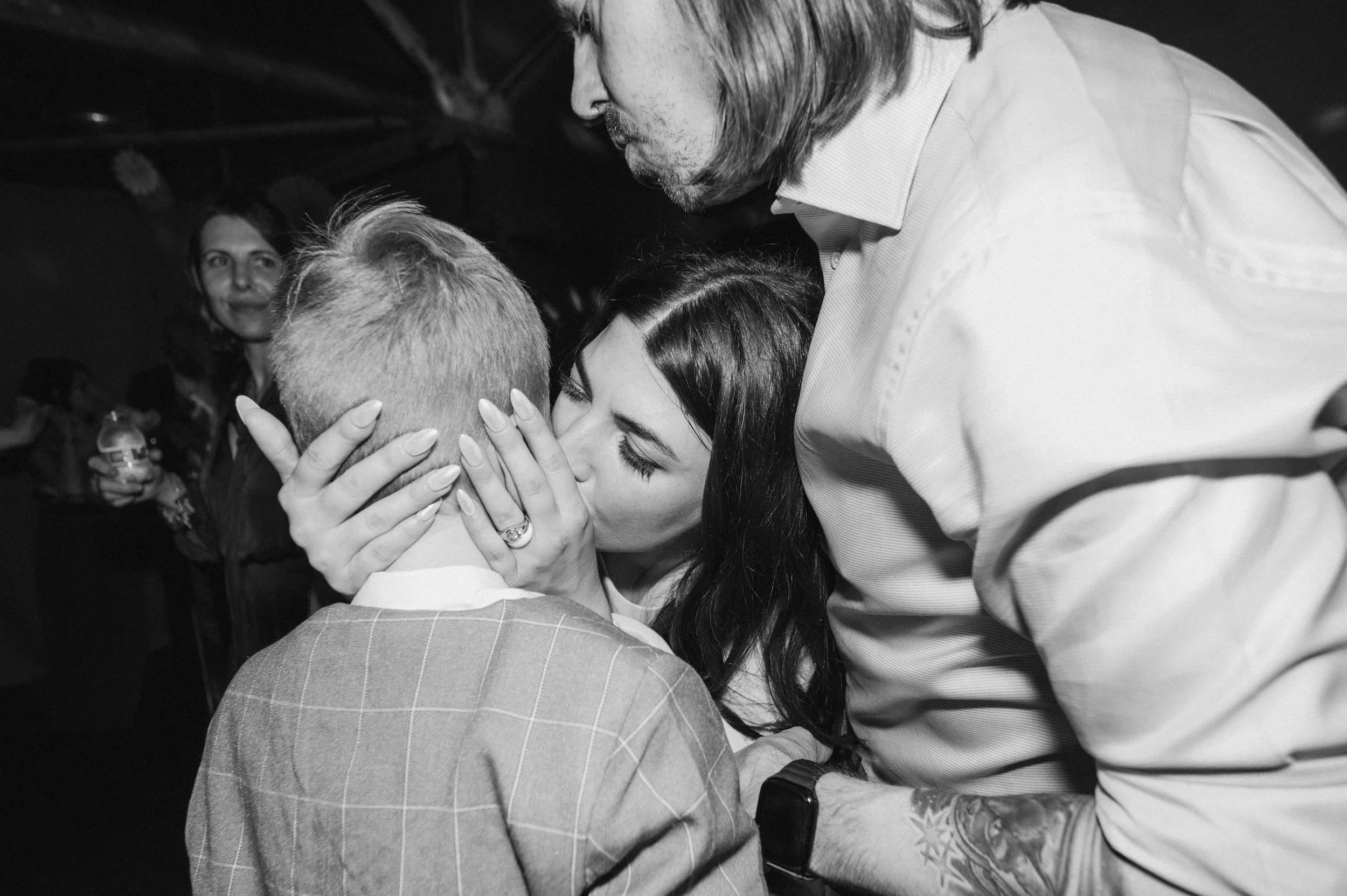 A woman kisses a man on the lips while holding his face. The woman has dark hair and is wearing a ring. The man has short light hair and is wearing a checked blazer. A man in a white shirt with a tattooed arm is leaning over them. In the background, 