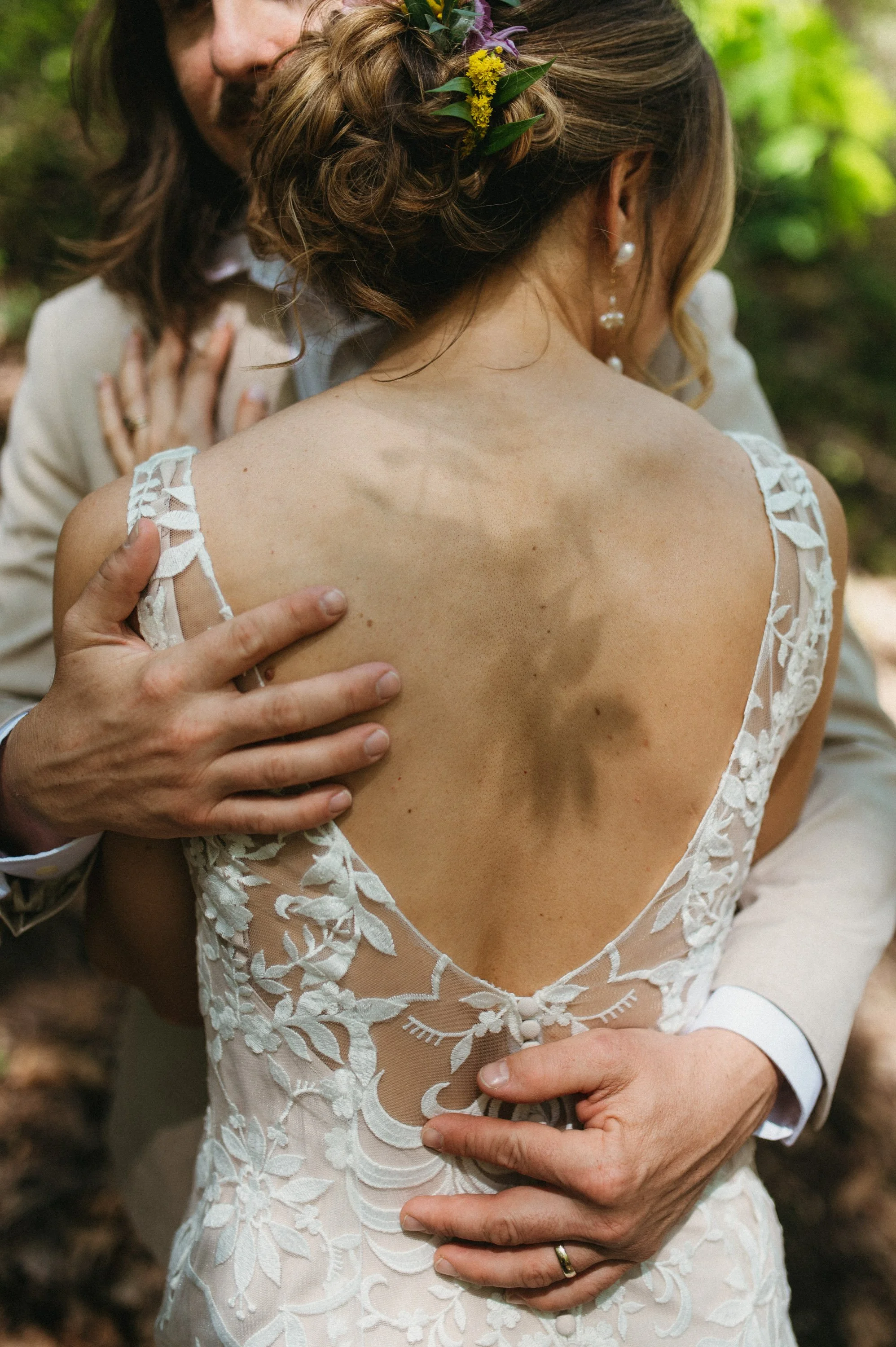 Close-up of a bride and groom embracing outdoors, focusing on the back of the bride's lace wedding dress and the groom's hand on her back.