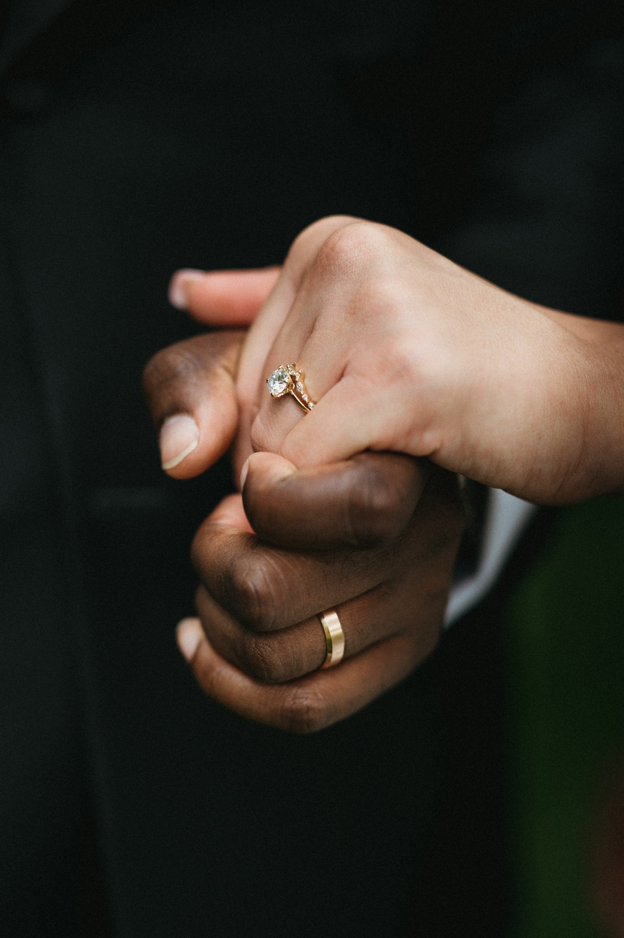 Close-up of a couple holding hands, showcasing their wedding rings. The woman has a diamond engagement ring on her finger, and the man has a simple gold wedding band.