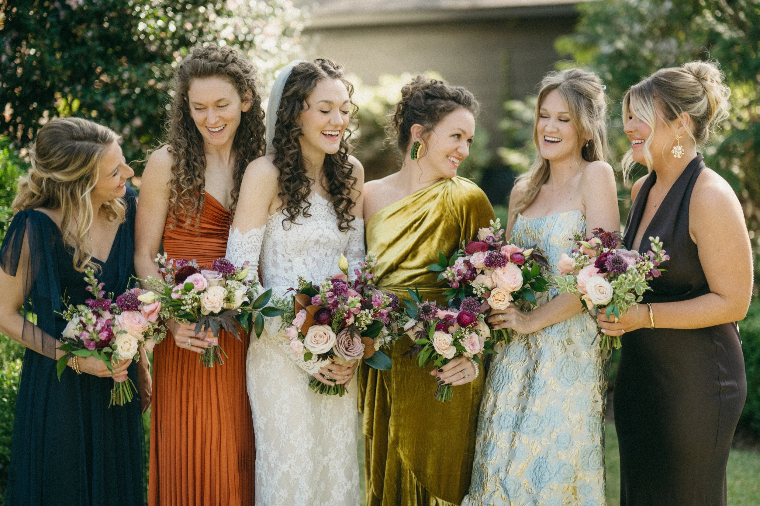 Seven women in elegant dresses holding bouquets of flowers, standing outdoors and smiling.