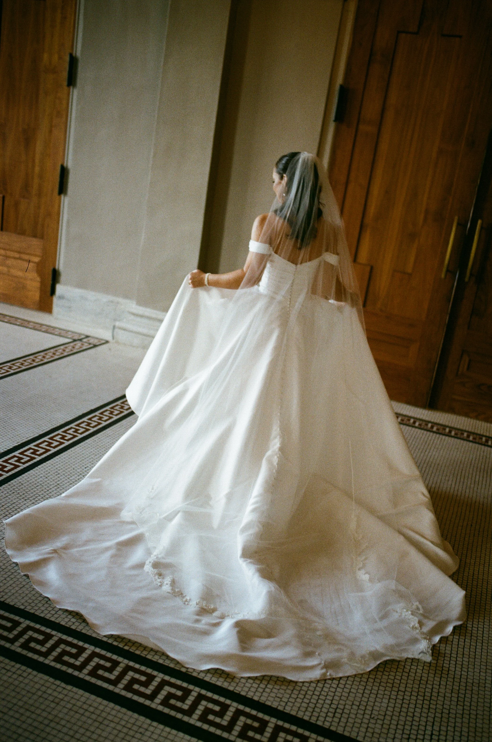 A bride in a white wedding dress and veil walking on a carpeted floor, holding the skirt of her dress. She is surrounded by wooden doors and a tiled floor.