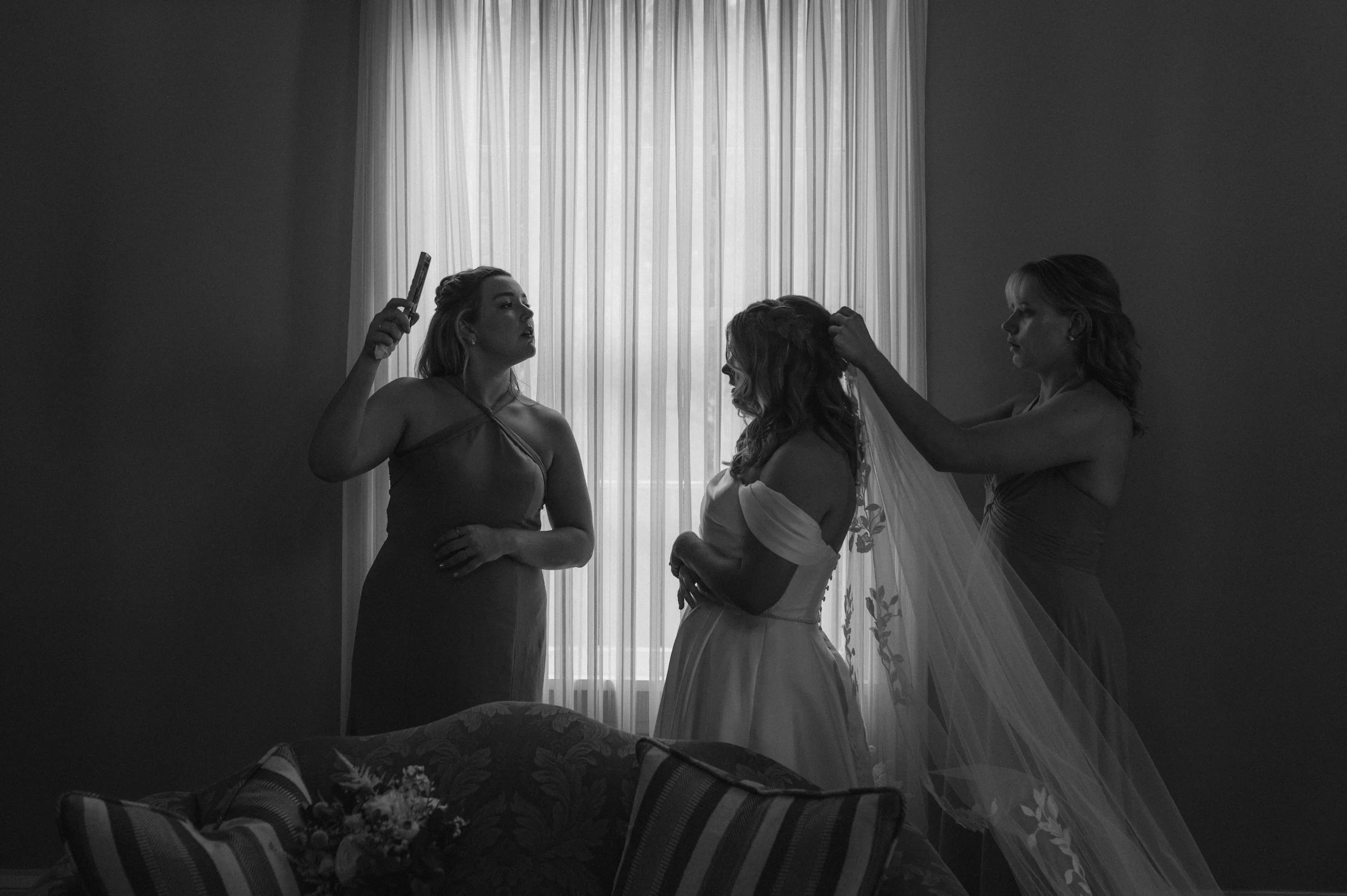 Three women in a room with a window and curtains; one woman in a wedding dress is having her hair done by another woman, while the third woman appears to be giving instructions, holding a smartphone in her hand.