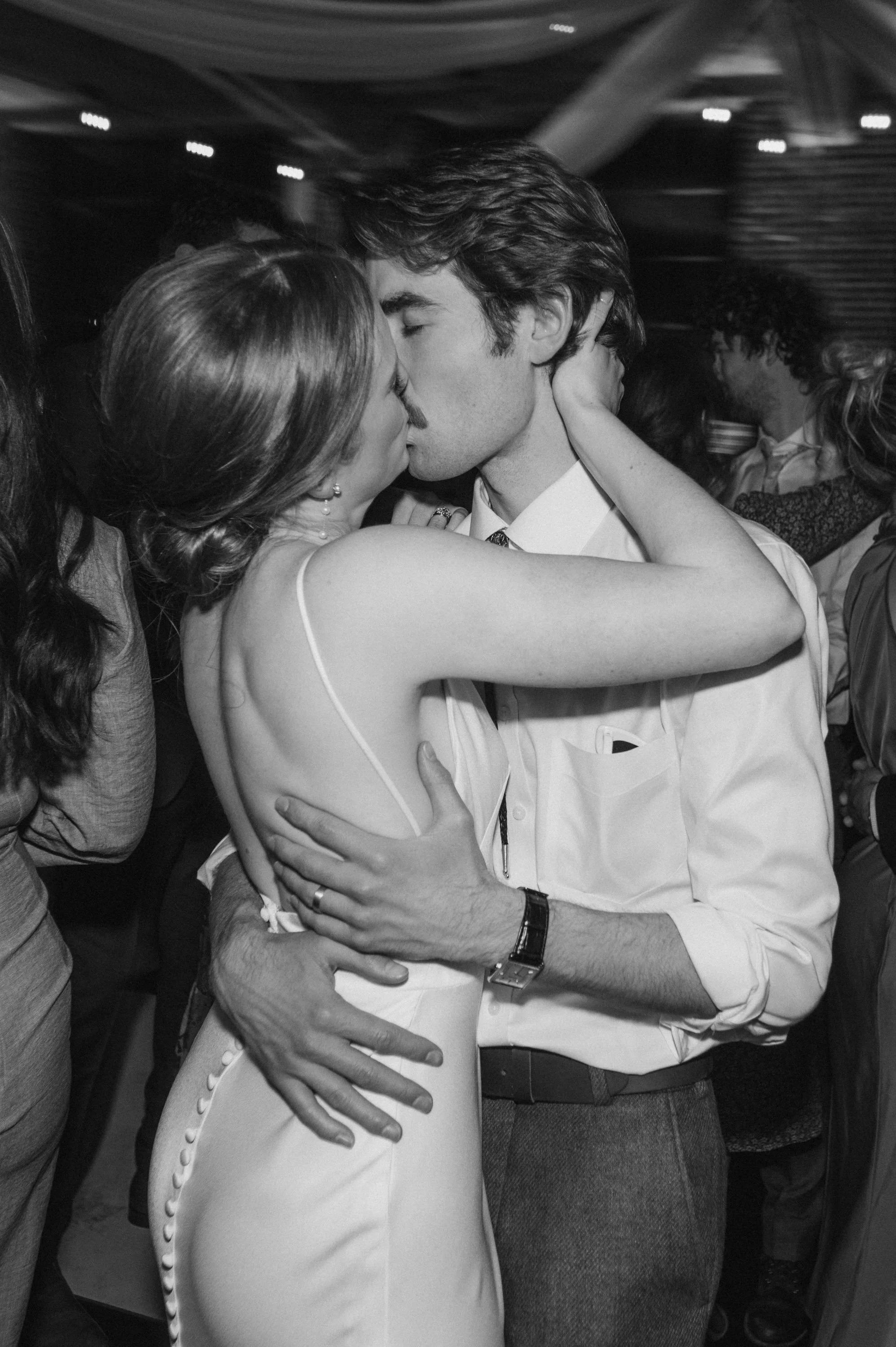 A man and woman kissing at a social gathering, with other people in the background.