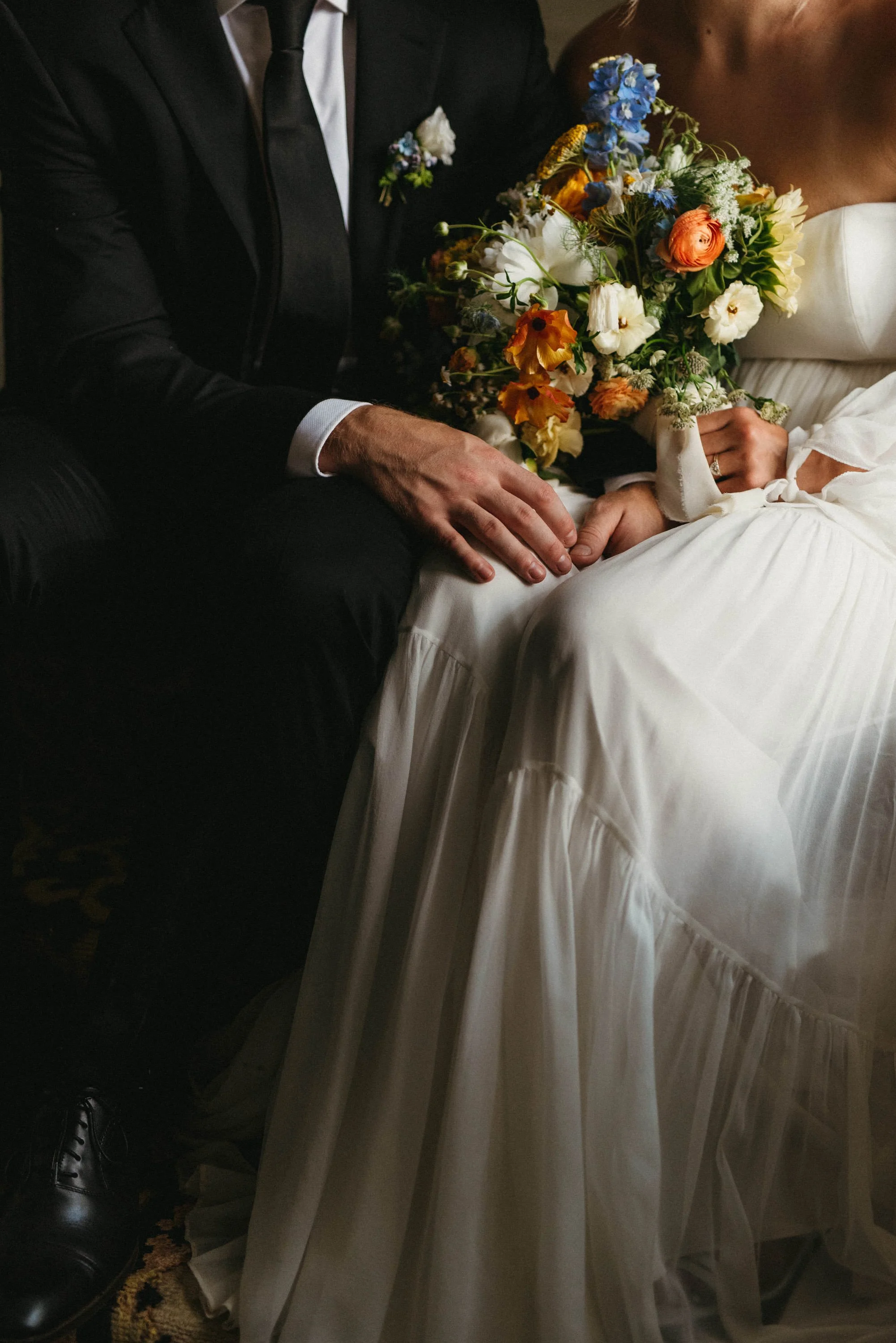 Close-up of a couple sitting closely, with the man's hand on the woman's lap. The woman is holding a vibrant bouquet of flowers. The scene suggests a wedding or romantic event.