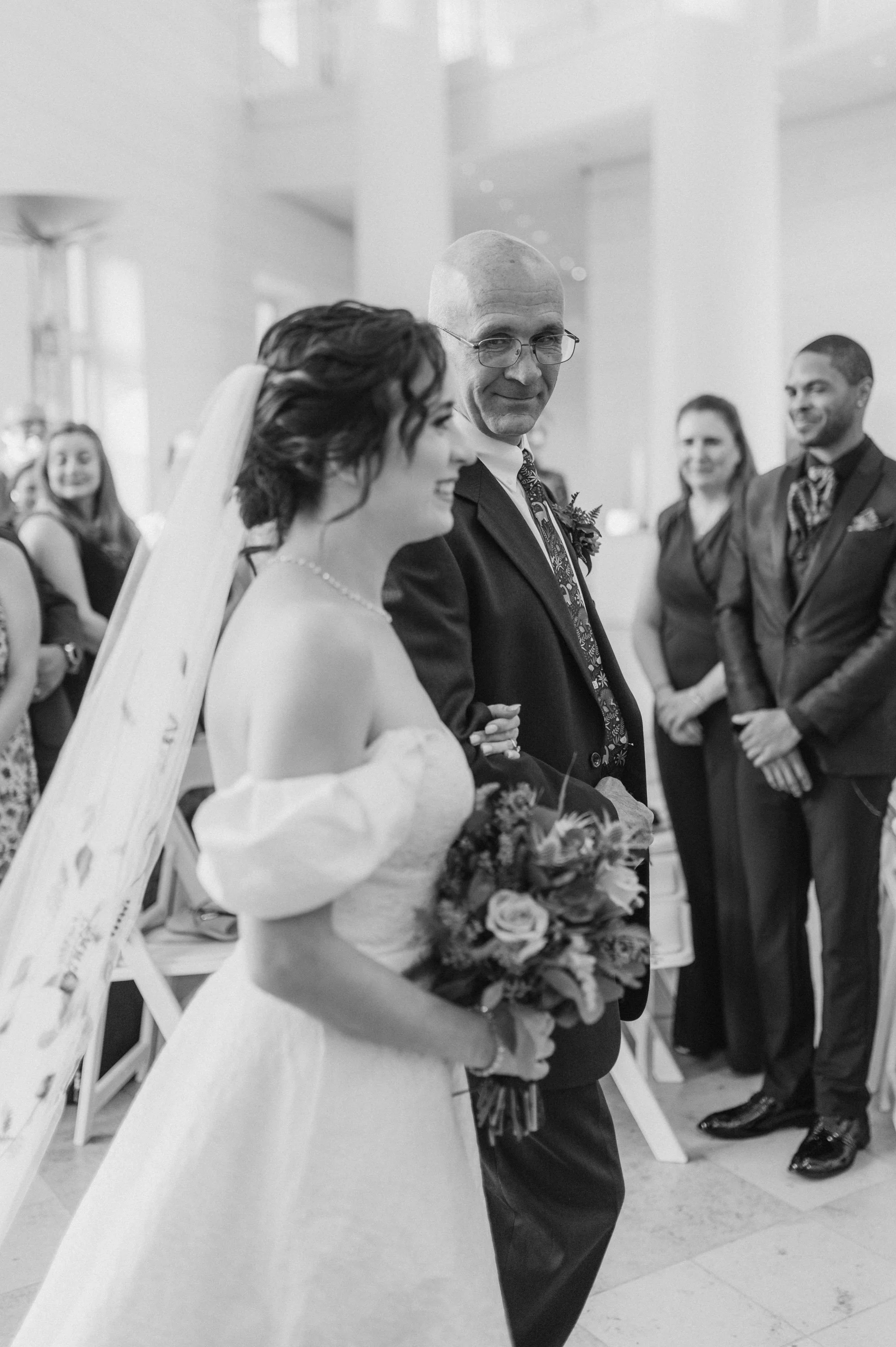 Black and white photo of a bride in a wedding gown holding a bouquet, walking down the aisle with a man. Guests are smiling and watching in the background.