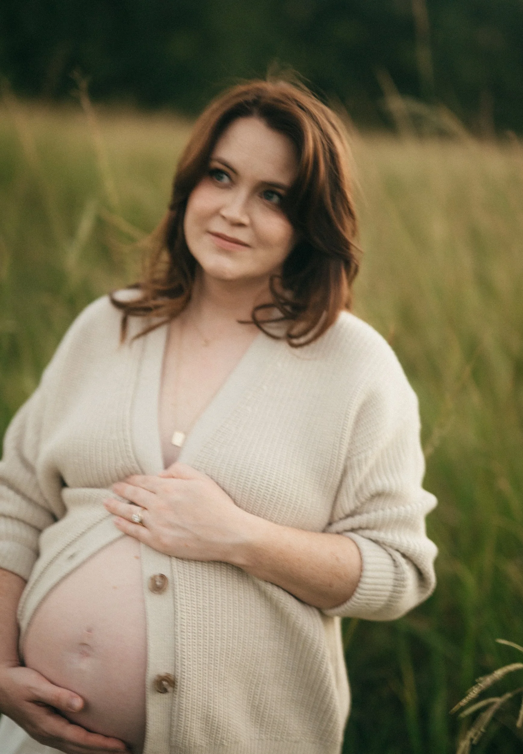 A pregnant woman with brown hair in a cream-colored cardigan stands outdoors in a grassy field during sunset, looking thoughtfully into the distance.