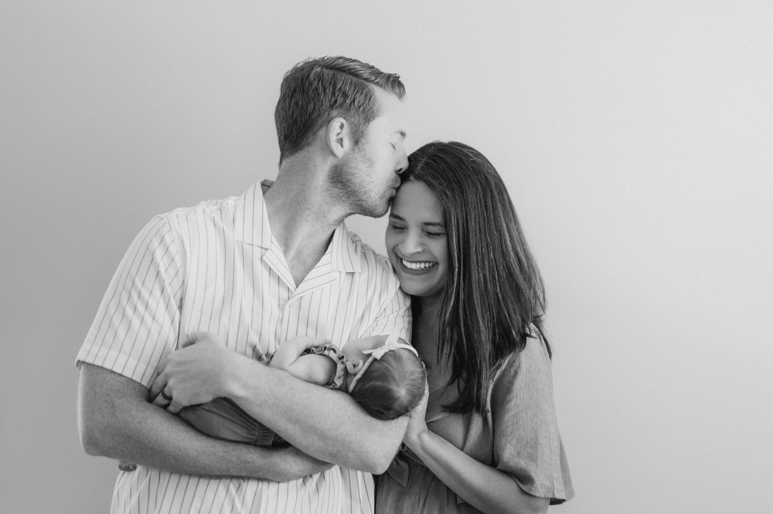 A man and woman holding a newborn baby, with the man kissing the woman's forehead, and both smiling. The photo is in black and white.