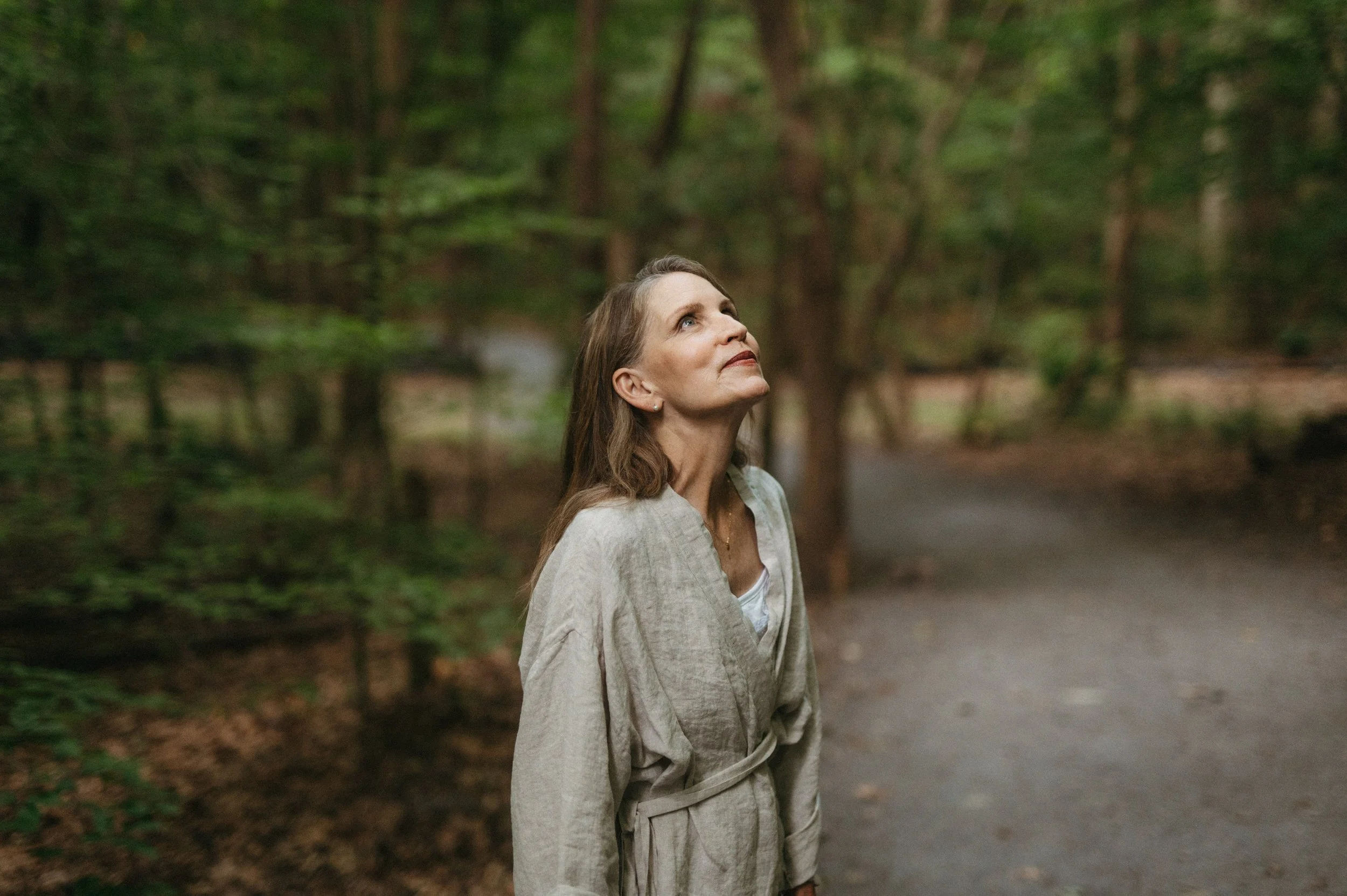 A woman in a light-colored robe stands on a forest trail, looking up thoughtfully at the trees surrounding her.