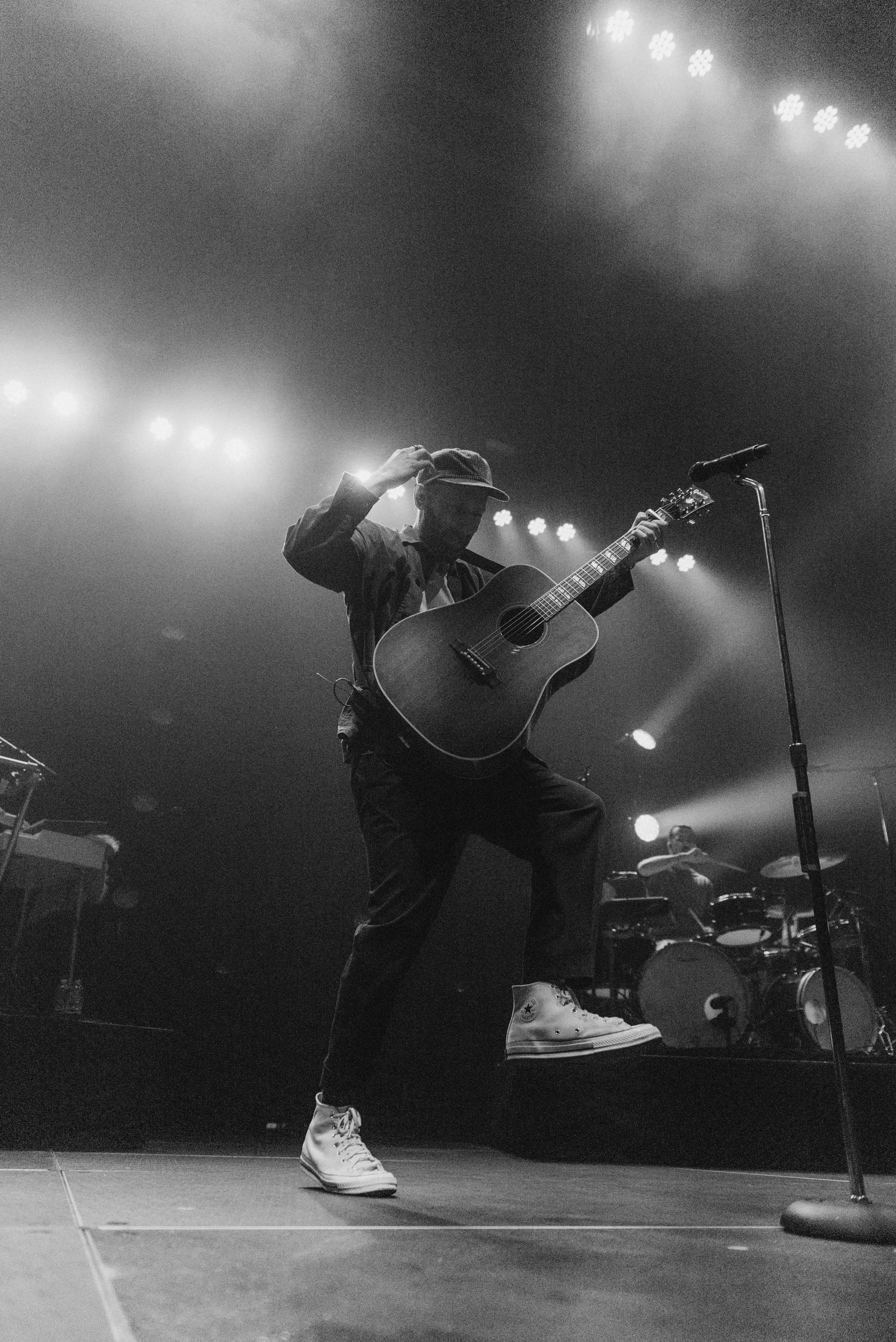 A musician on stage playing an acoustic guitar, wearing sneakers, a hat, and stylish clothing, with stage lights shining overhead.