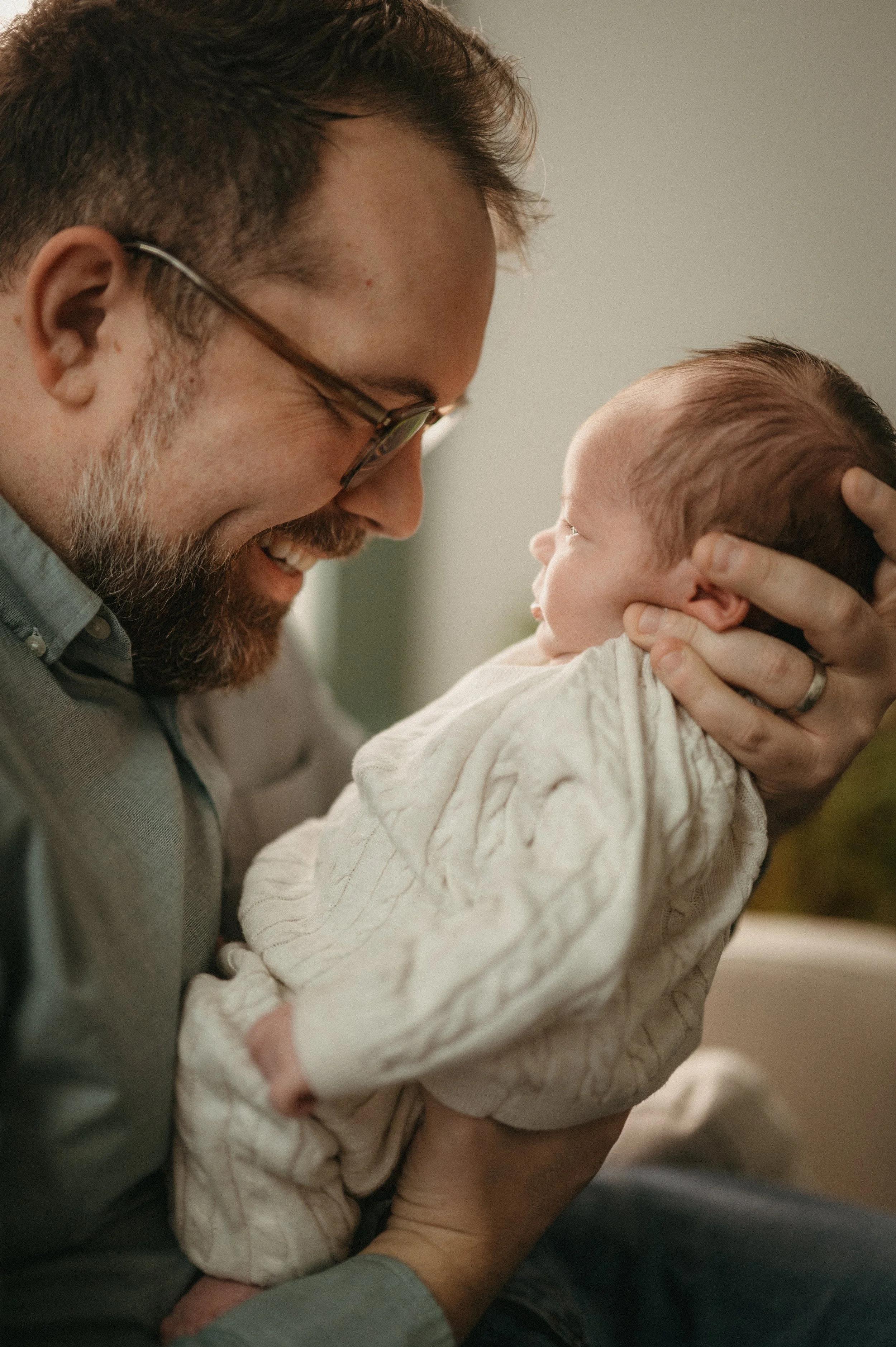 A man with glasses and a beard holding a baby dressed in a cream-colored sweater, smiling and leaning close to the baby indoors.