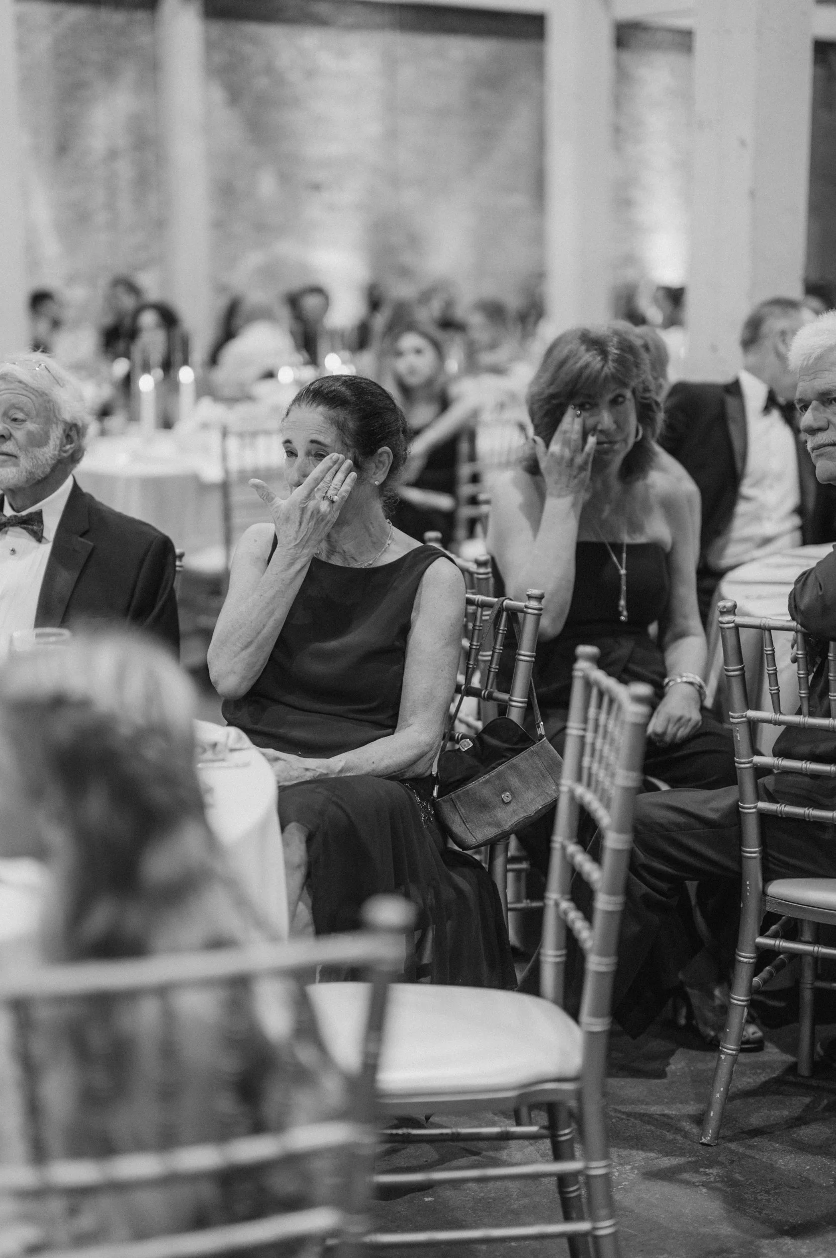 Black and white photo of women at a formal event, some wiping tears from their eyes, seated around round tables with chairs.