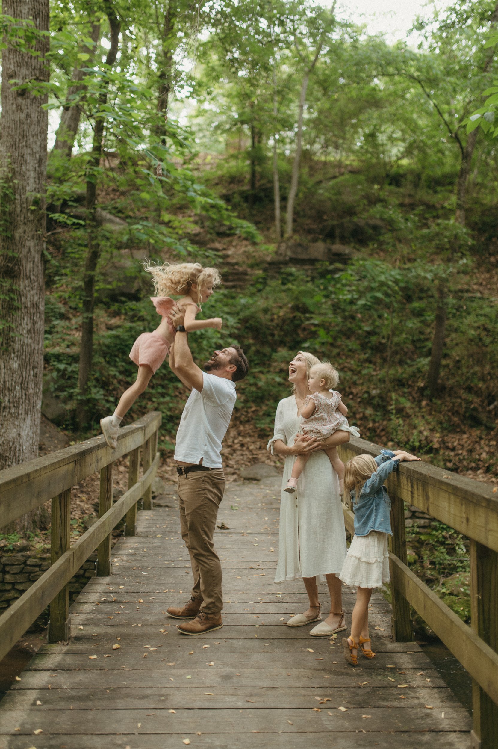 A family of five stands together on a path in the woods, the parents kiss while the children look around