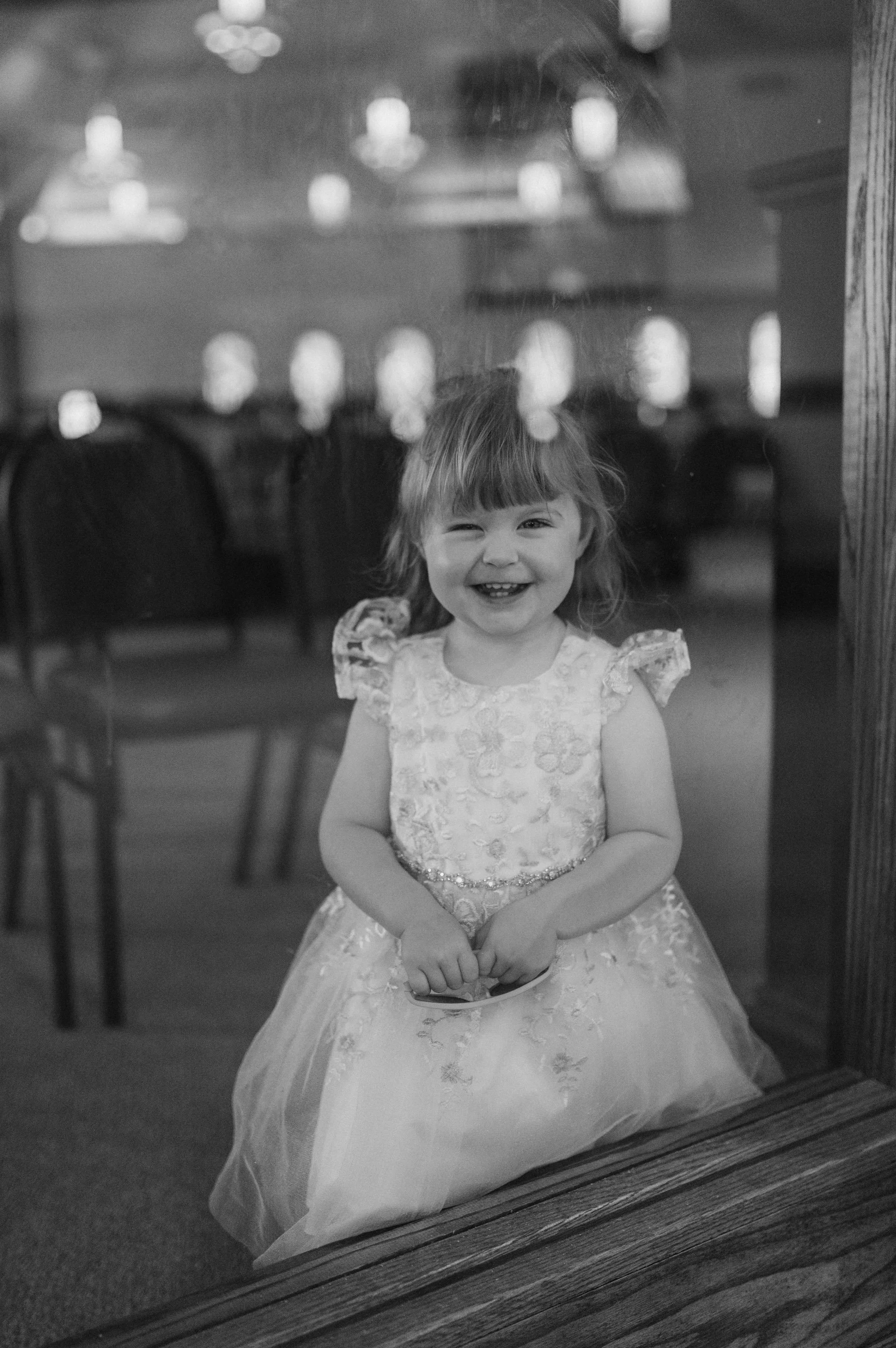 A young girl in a fancy dress smiling and winking, seen through a glass window, with chairs and lights in the background.