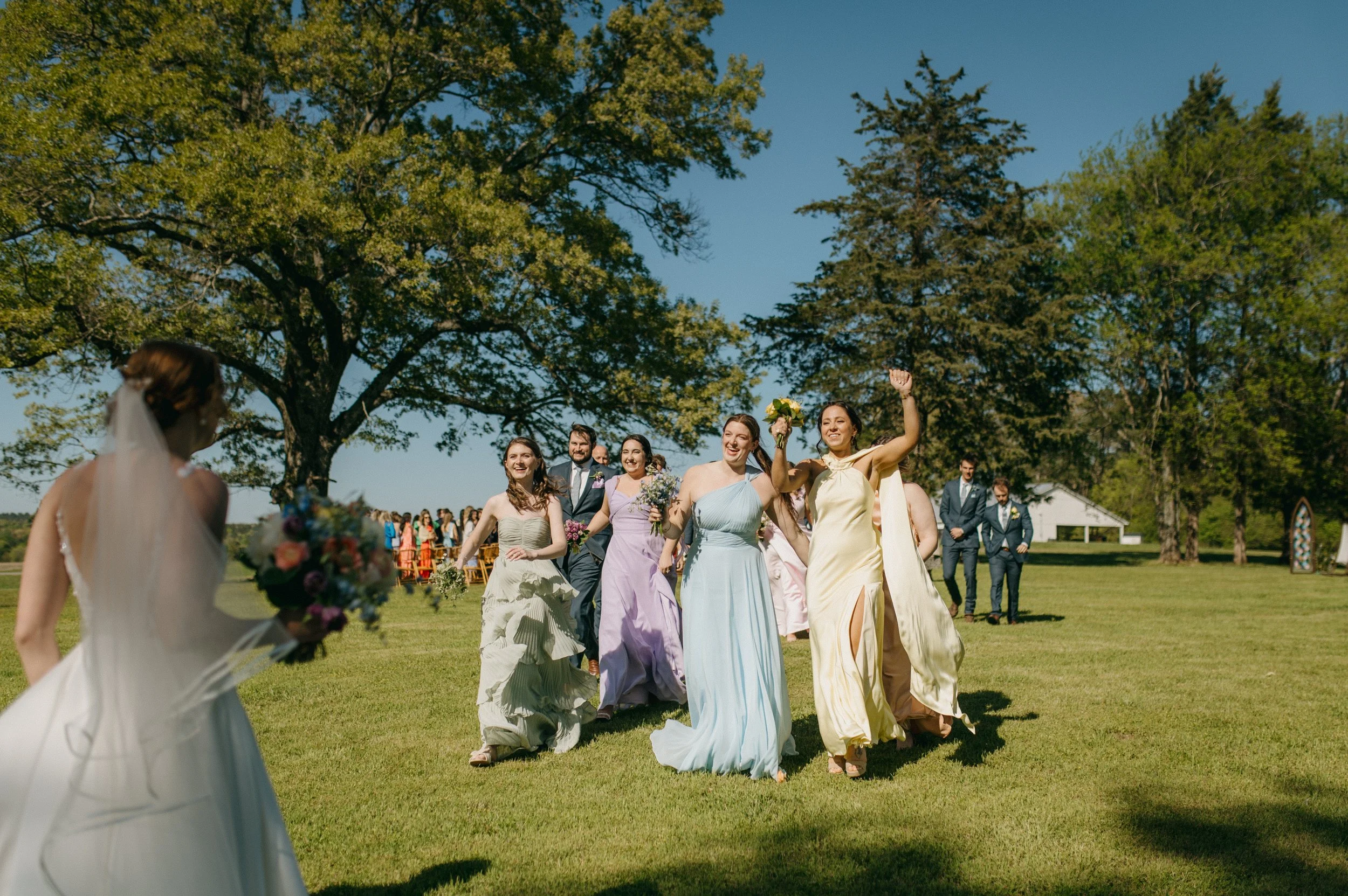 Group of wedding guests running towards bride who is holding a bouquet outdoors on a sunny day, with trees in the background.
