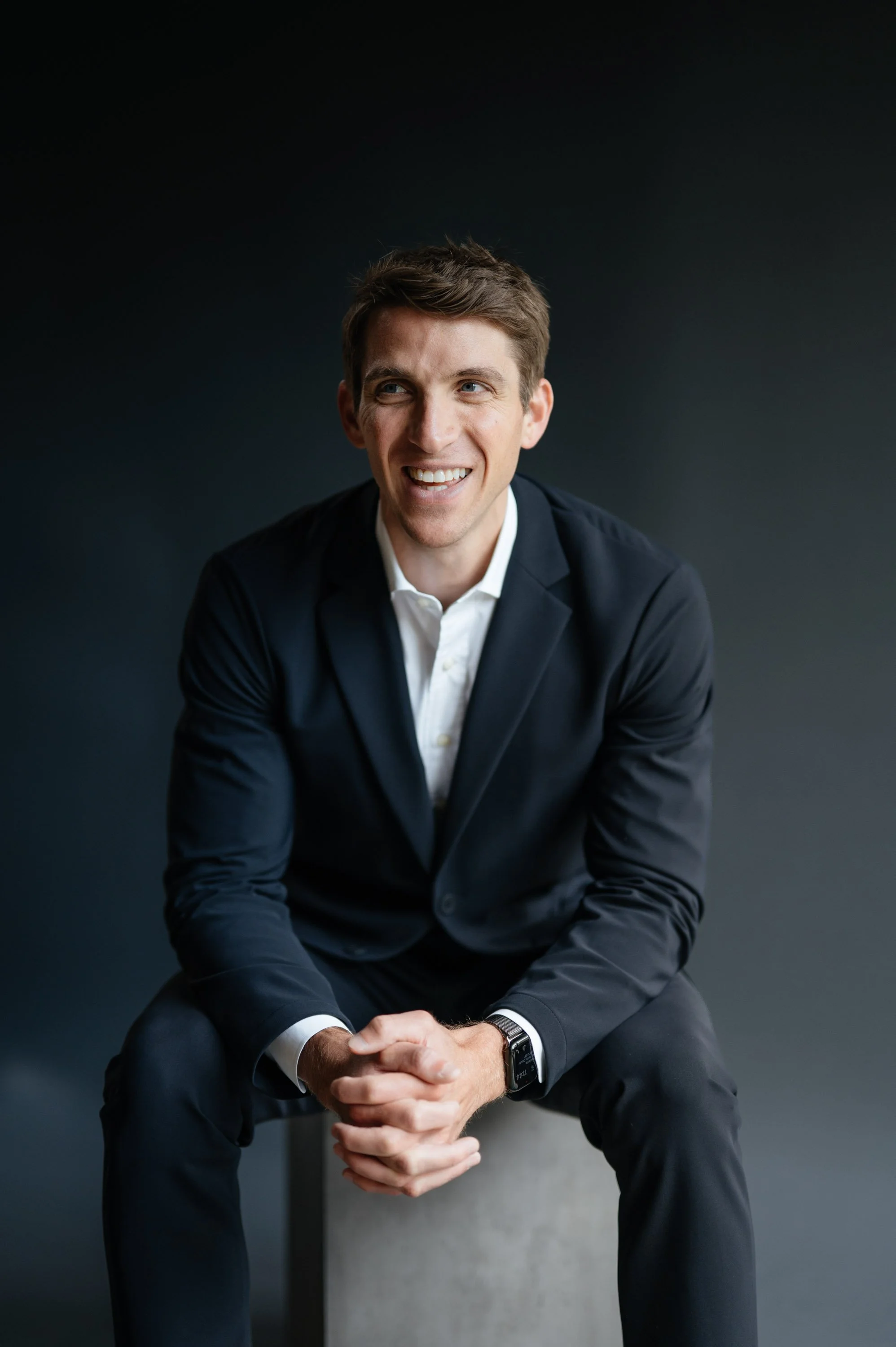 A man smiling and sitting on a white cube against a dark background, wearing a black suit and a white shirt.