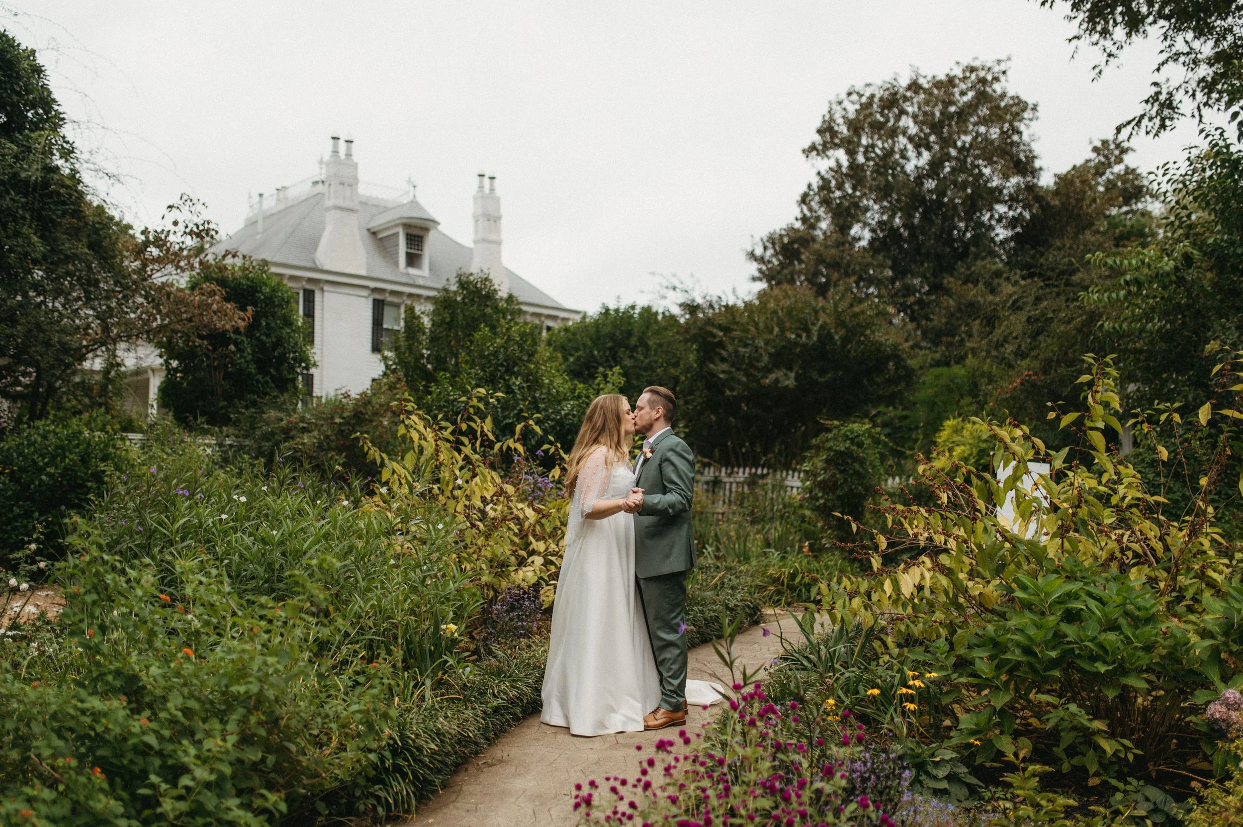 A bride and groom kissing in a lush garden with a large white house in the background.