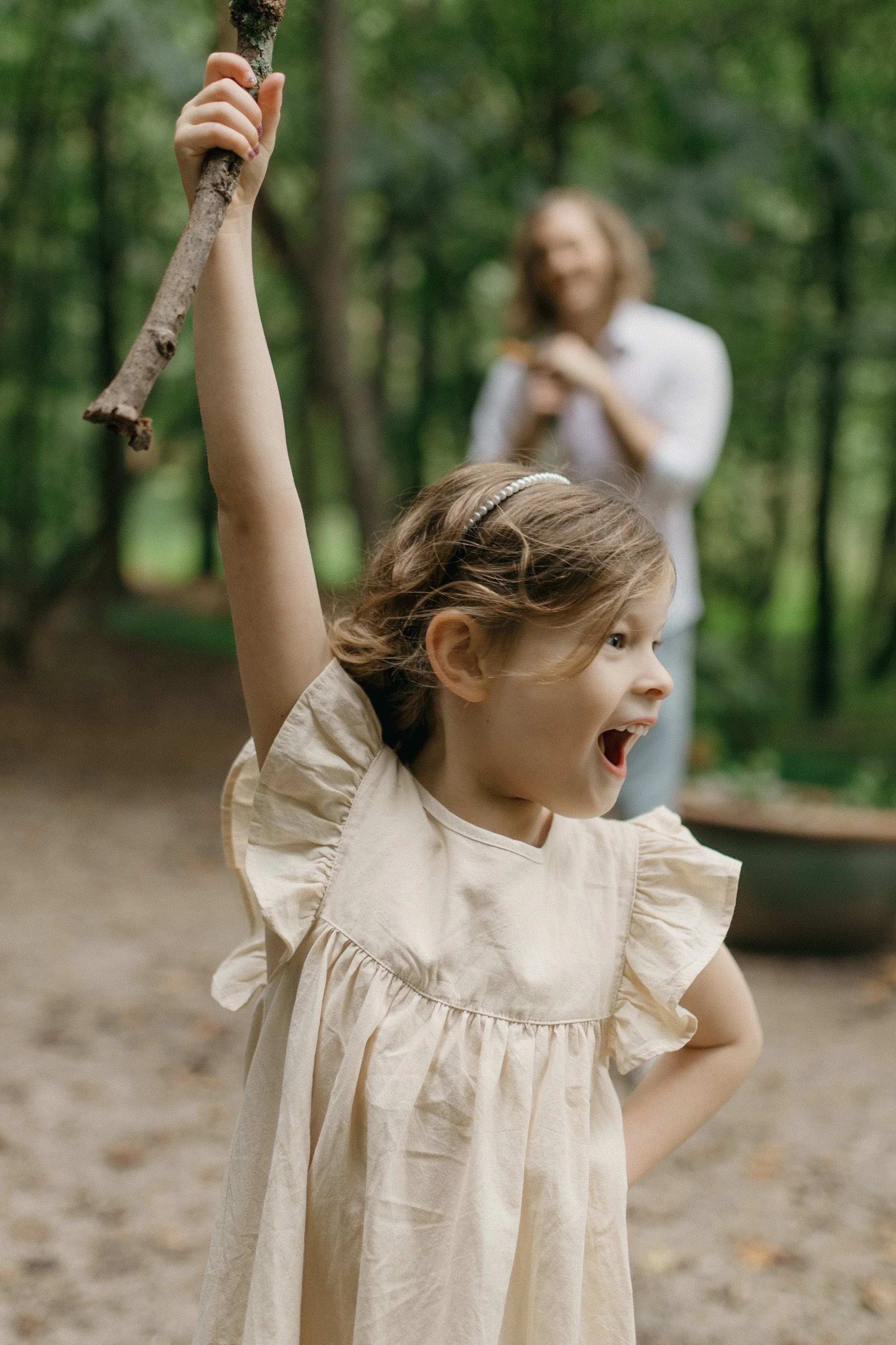 A young girl with a headband holding a stick, smiling with her mouth open and one hand on her hip, outdoors in a wooded area, with a man blurred in the background.