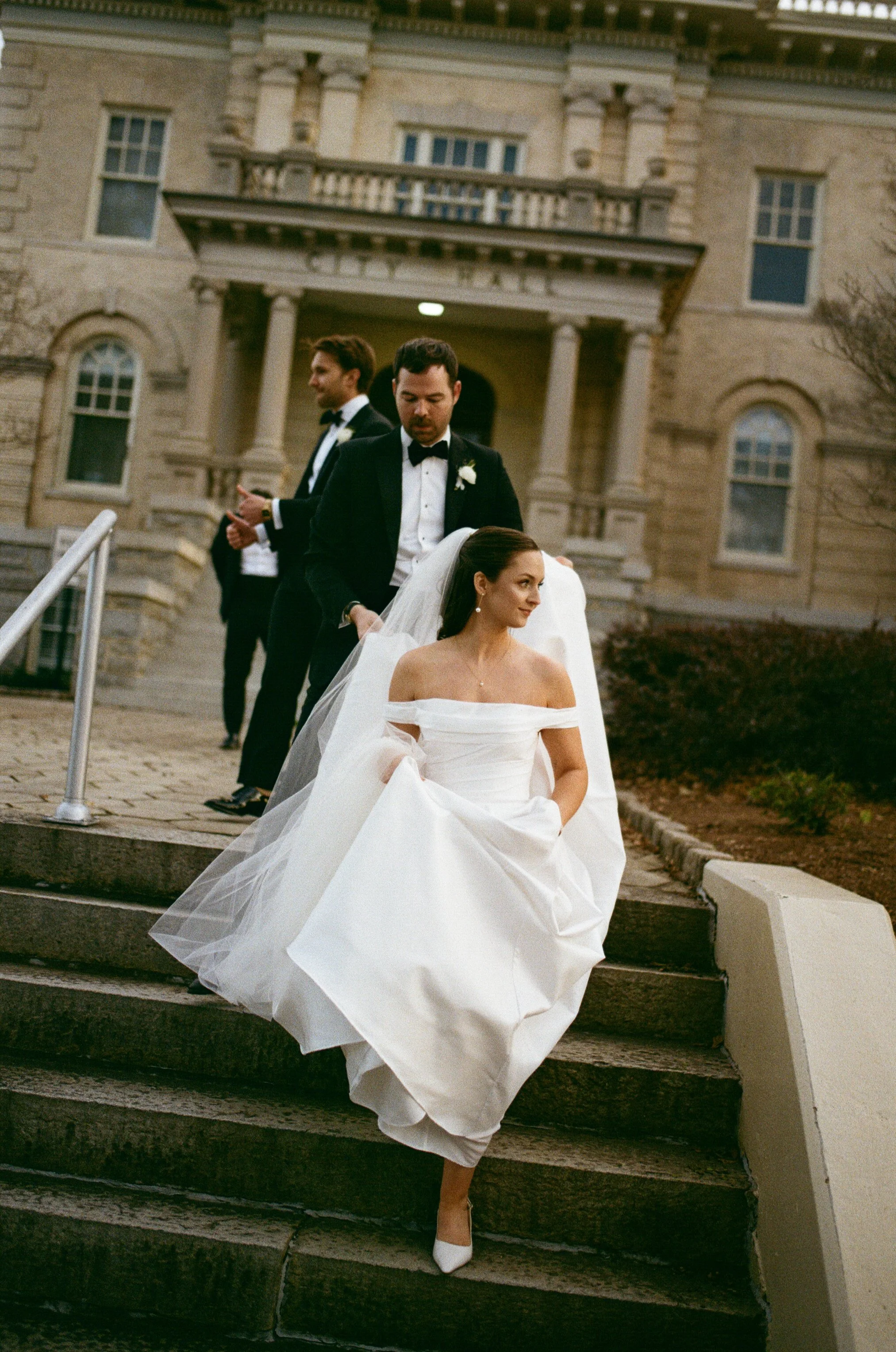 Bride in an off-shoulder white wedding gown and veil sitting on outdoor stone steps, with three men in tuxedos ascending behind her, in front of a large historic building.