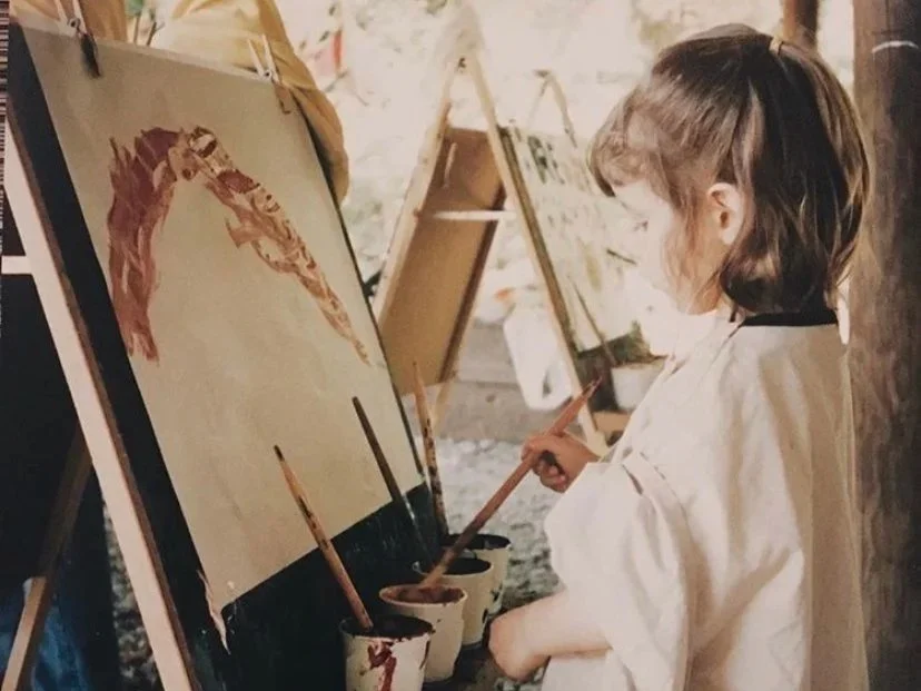 A young girl in a white smock painting a horse portrait on a canvas in an art studio.